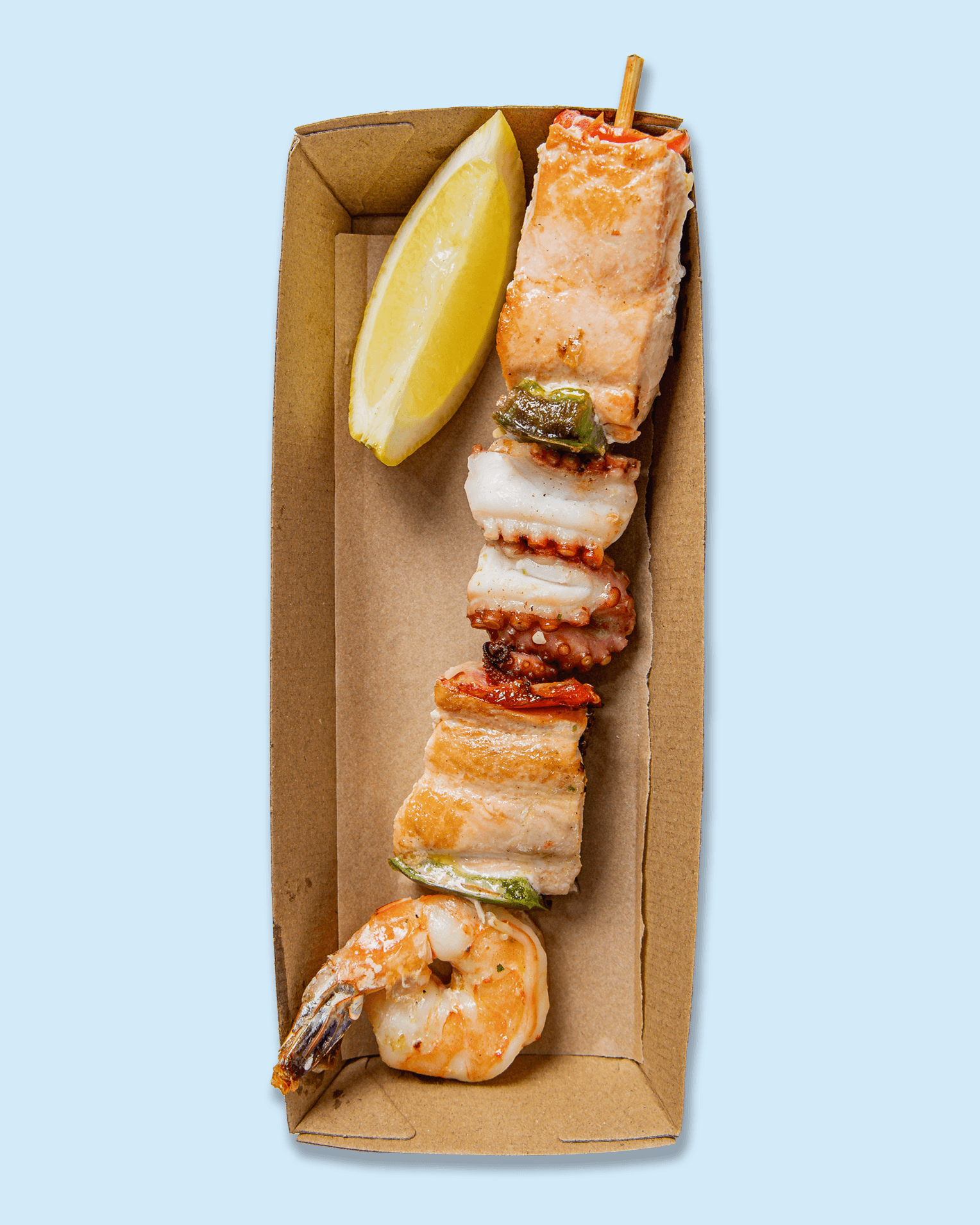 A cardboard tray with grilled seafood skewers, lemon wedges, and a slice of lemon on specialized catering paper with 'Nicholas catering & events' logo repeated on it.