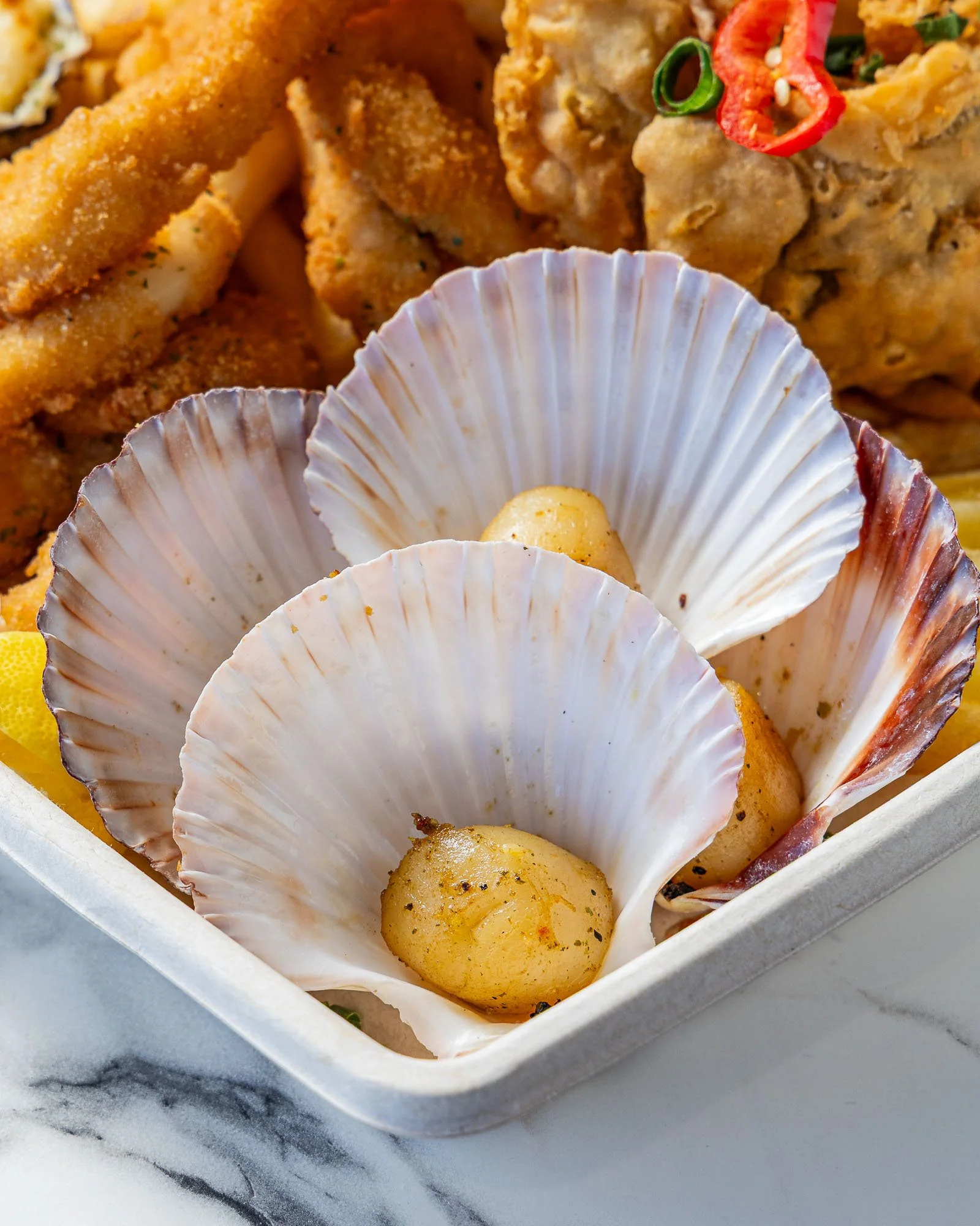 Close-up of baked scallops in shell with garnishes on top, inside a white container.