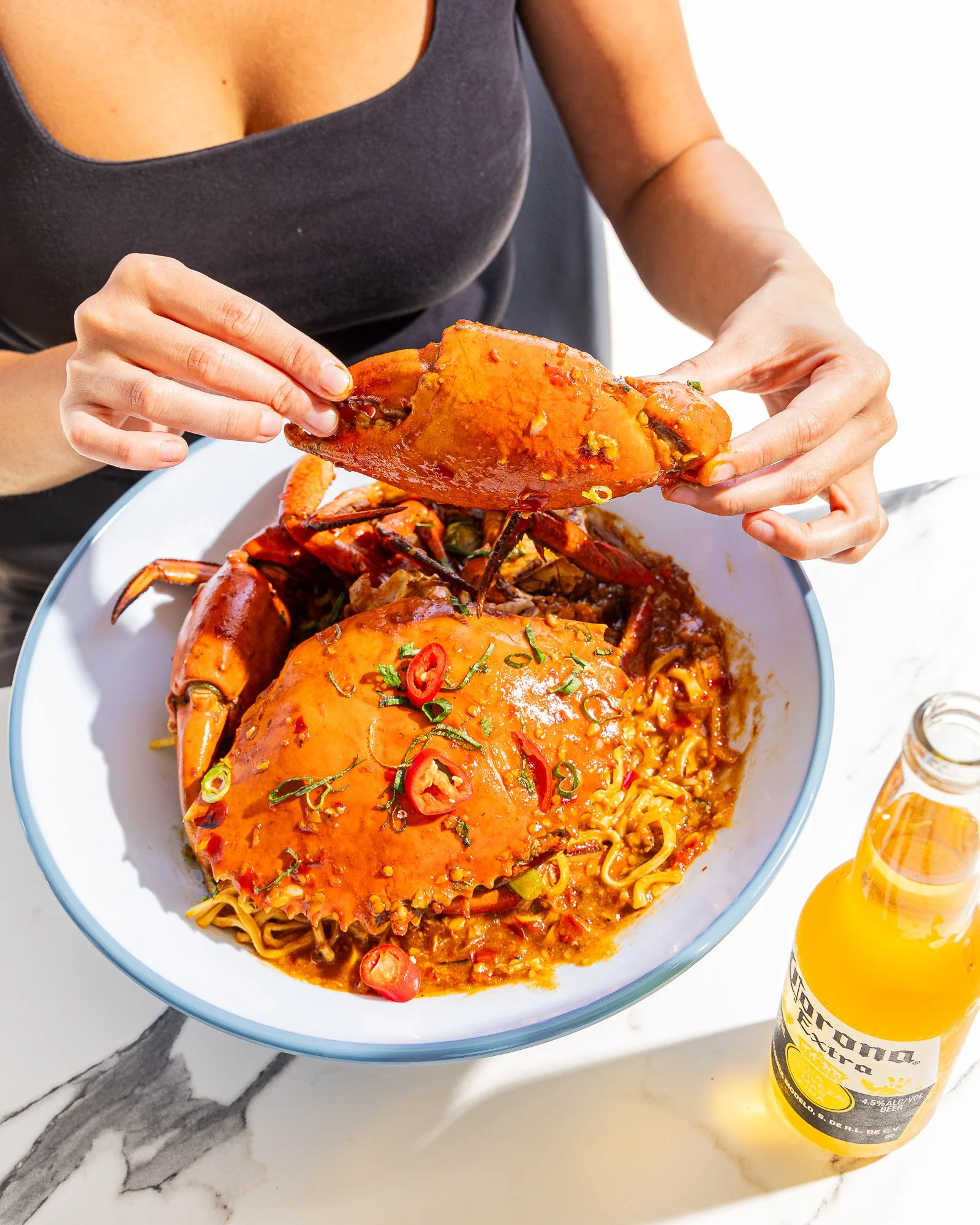 A person holding a crab leg over a bowl of noodles, garnished with sliced chili peppers and green onions. A bottle of Corona Extra beer is on the table next to the bowl.