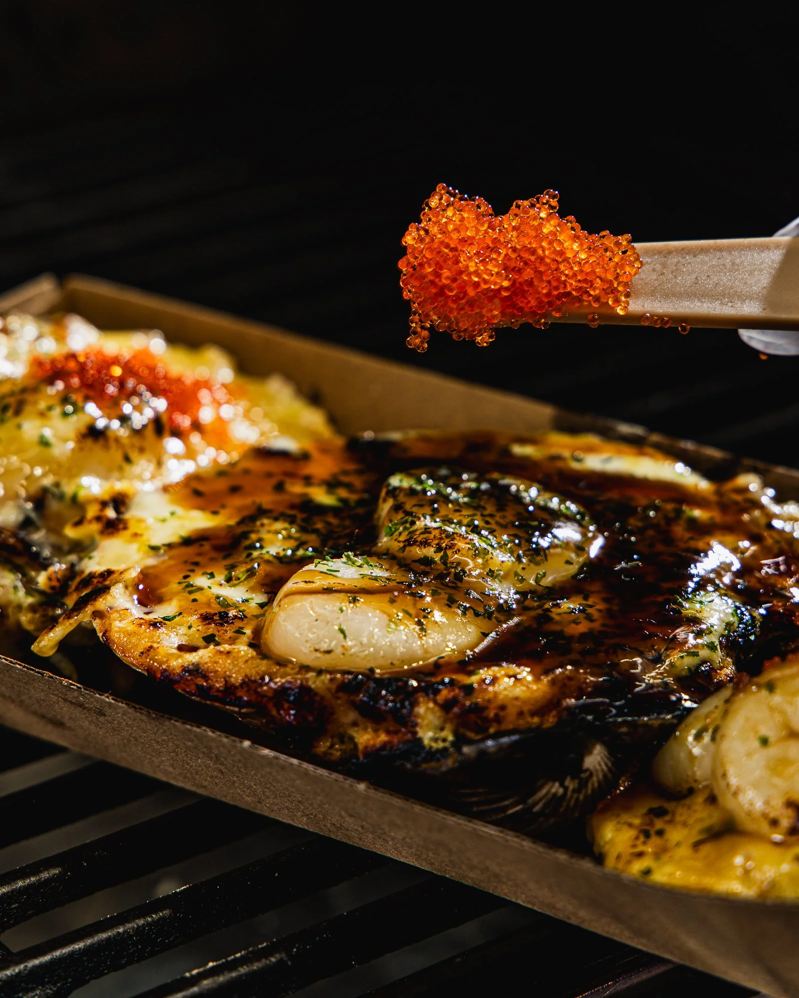 Close-up of a spatula with orange fish roe above cooked seafood, possibly calamari, on a grill.