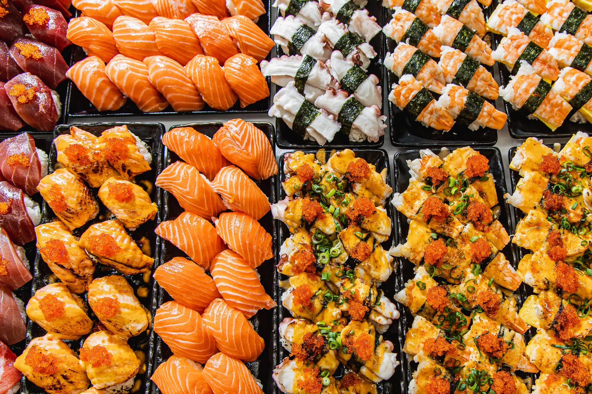 Assorted sushi rolls and sashimi, including salmon, tuna, and cooked sushi with garnishes, displayed on black trays.