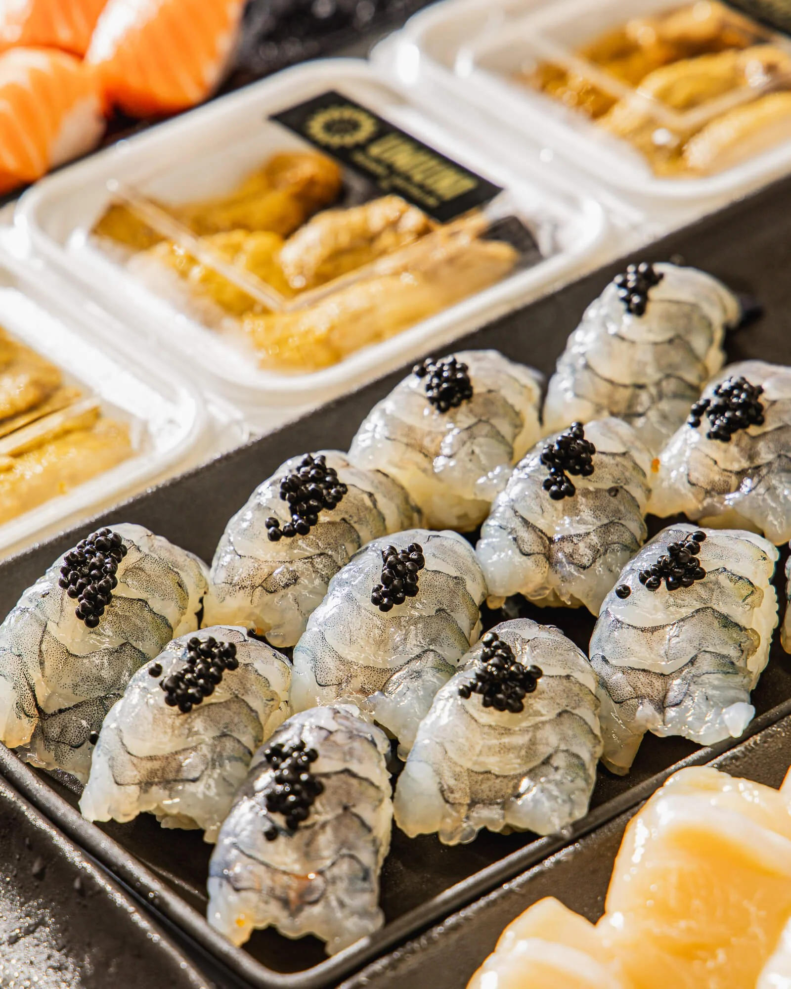 Close-up of a tray of sushi rolls topped with black caviar, with slices of salmon and other seafood in the background.