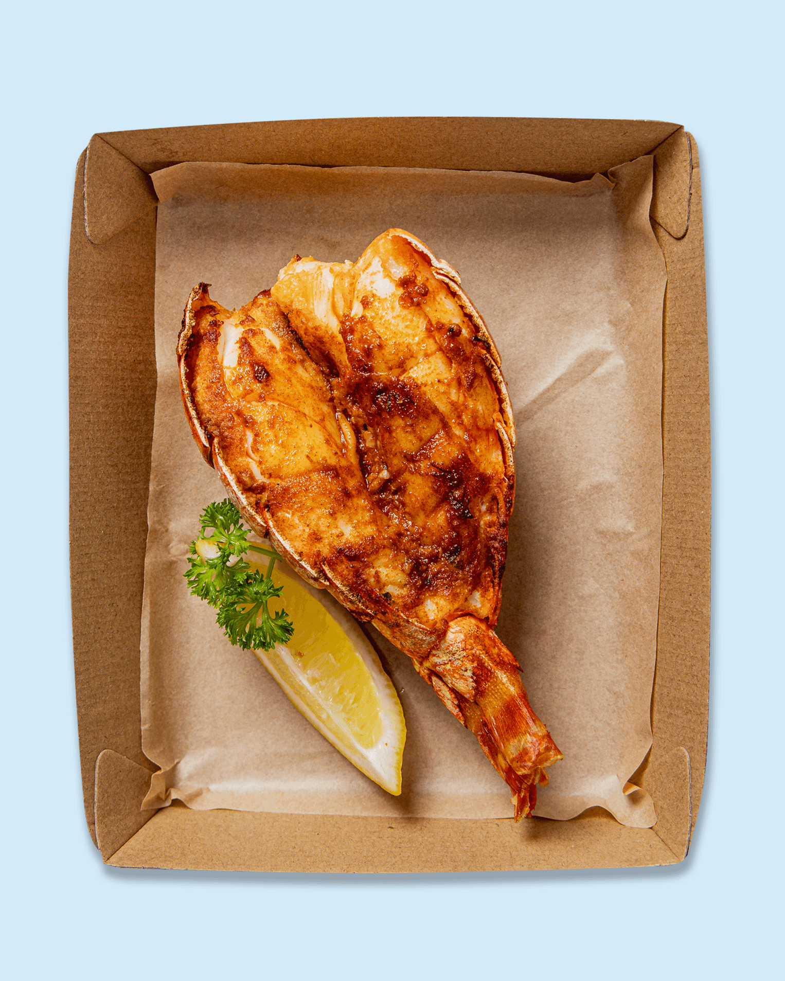 Lobster tail with grilled seasoning, lemon wedge, and parsley garnish in a brown box on a paper plate.