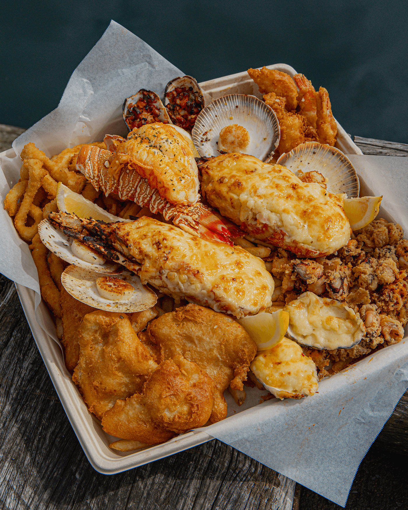 A takeout container filled with fried seafood, shrimp, fish, and crab cakes, garnished with lemon wedges and surrounded by shells and seafood.