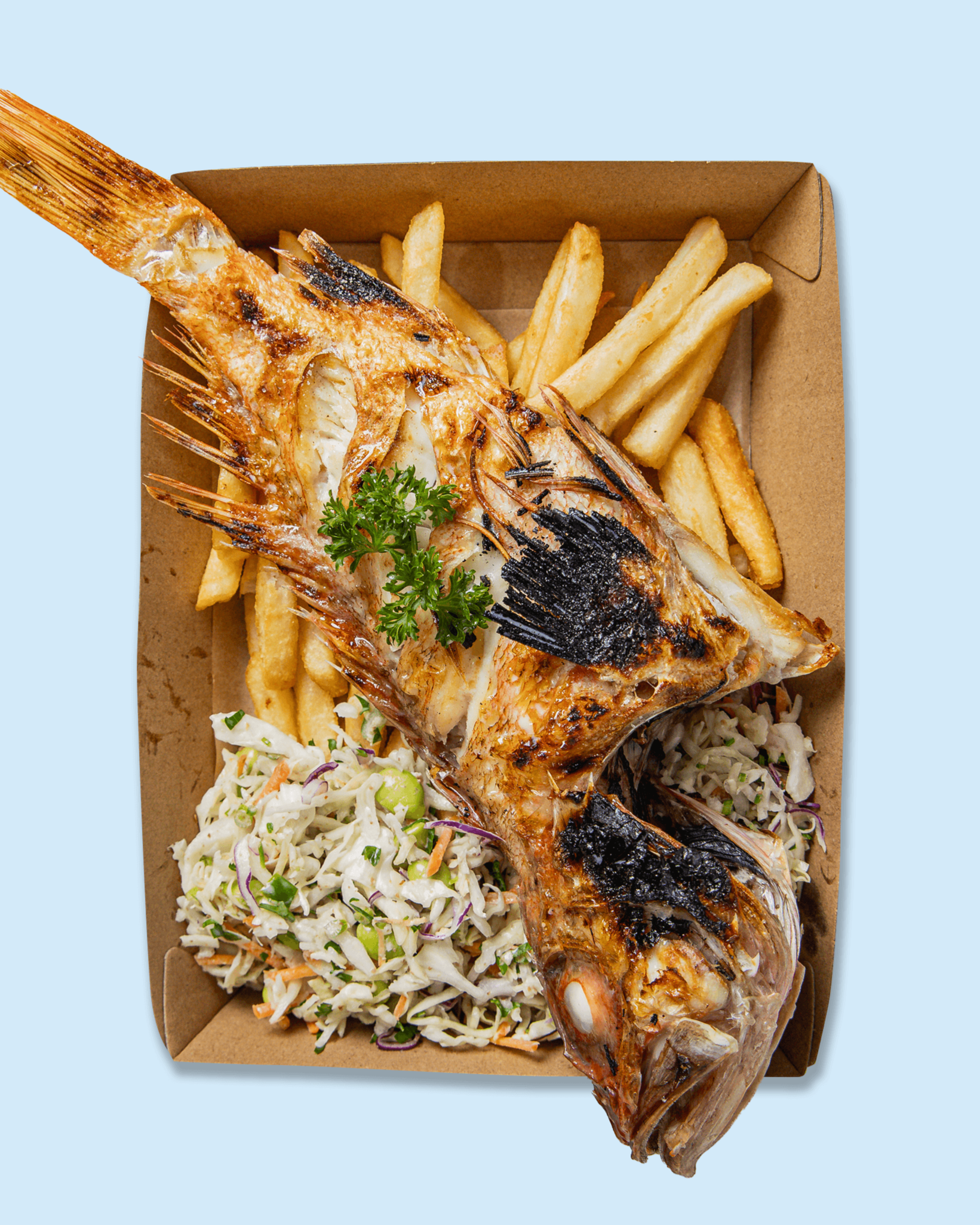 A whole grilled fish with crispy skin served on a bed of coleslaw, garnished with parsley, and accompanied by French fries and a lemon wedge in a cardboard tray.