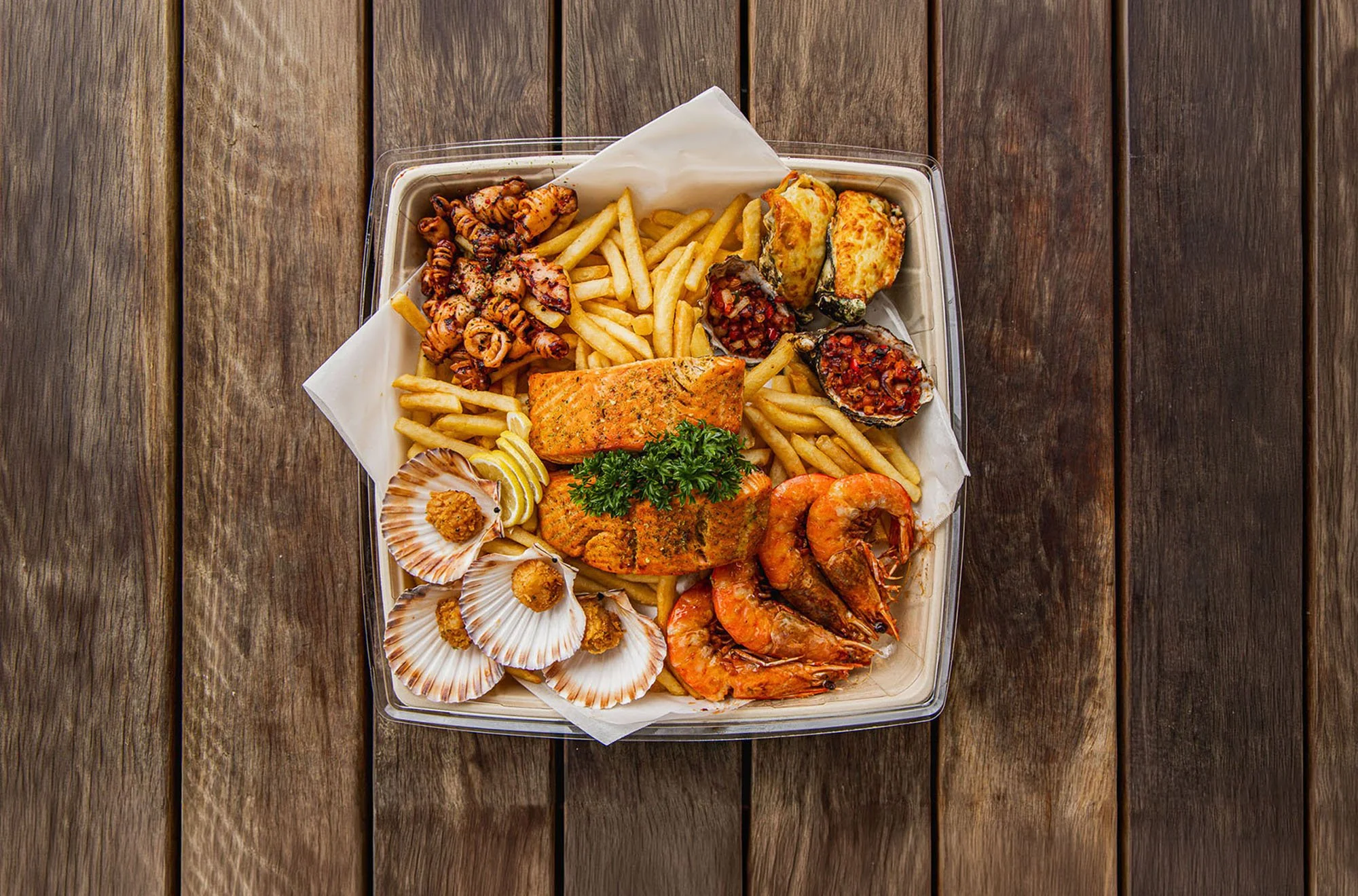 A takeout box filled with fried shrimp, battered fish, scallops, and various seafood dishes on a marble surface.