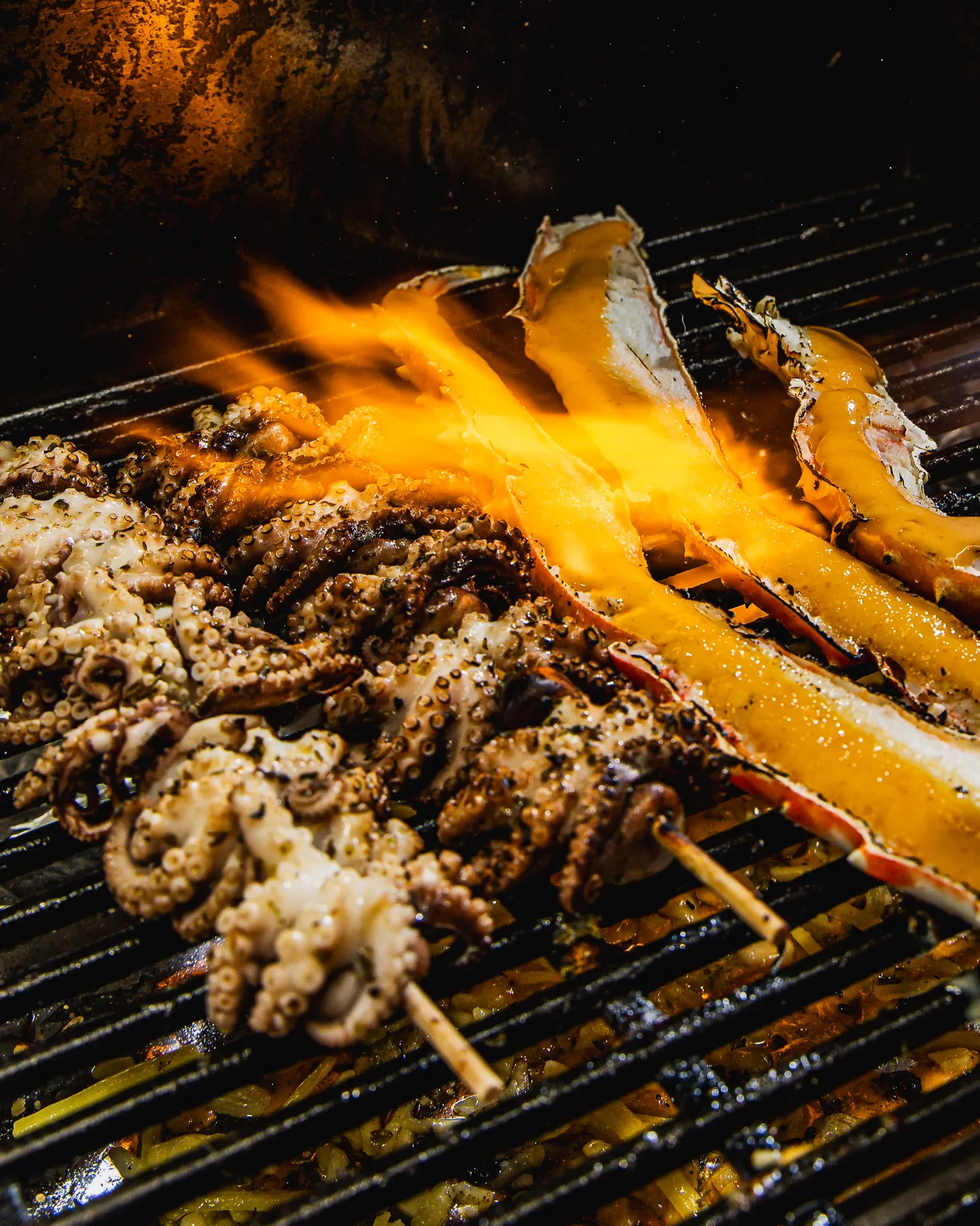 Seafood skewers, including octopus and fish, being grilled over an open flame with visible grill grates and glowing heat.