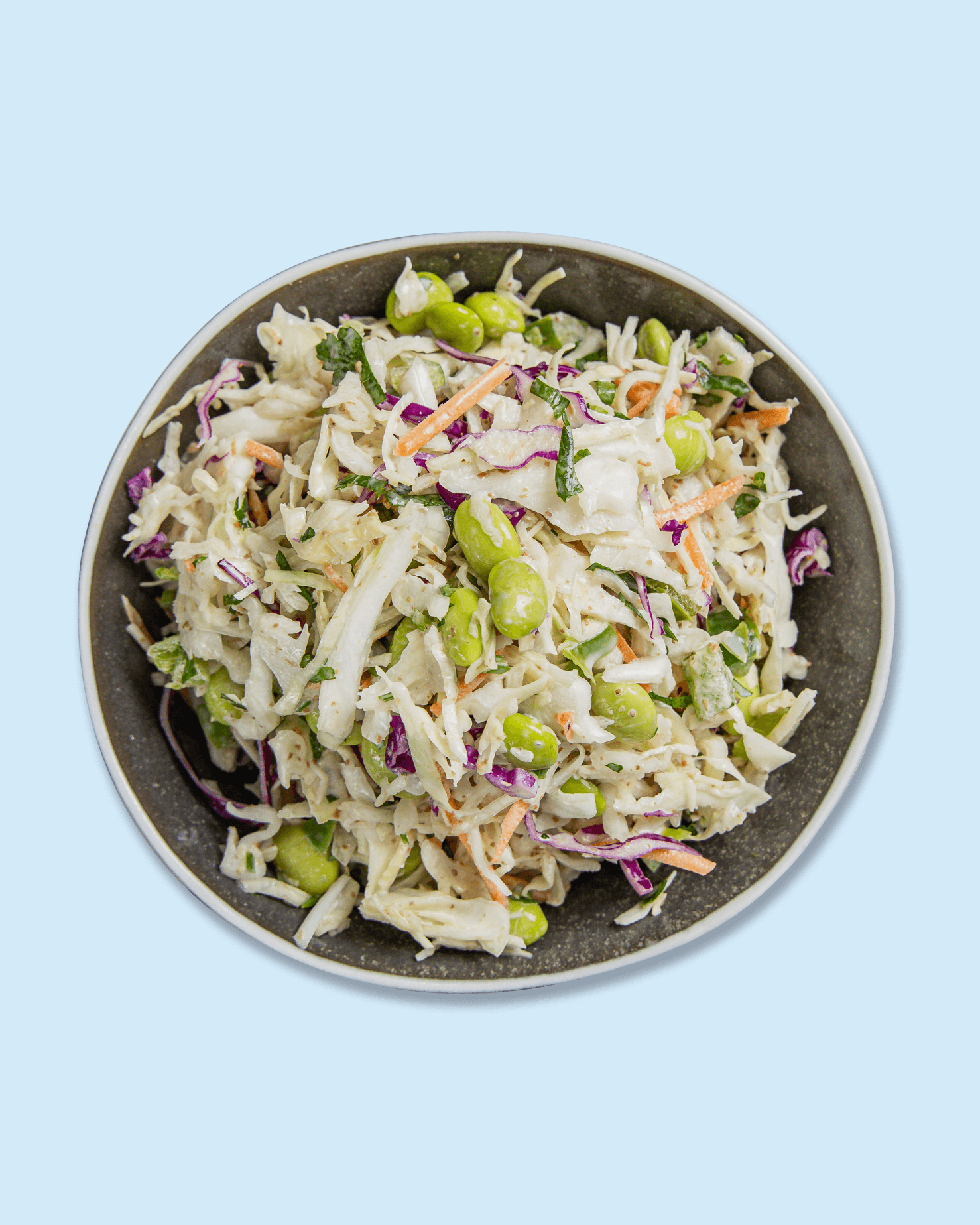 A bowl of coleslaw salad with shredded cabbage, carrots, green peas, and chopped herbs, placed on a white surface with a repeated 'Nicholas' logo pattern.