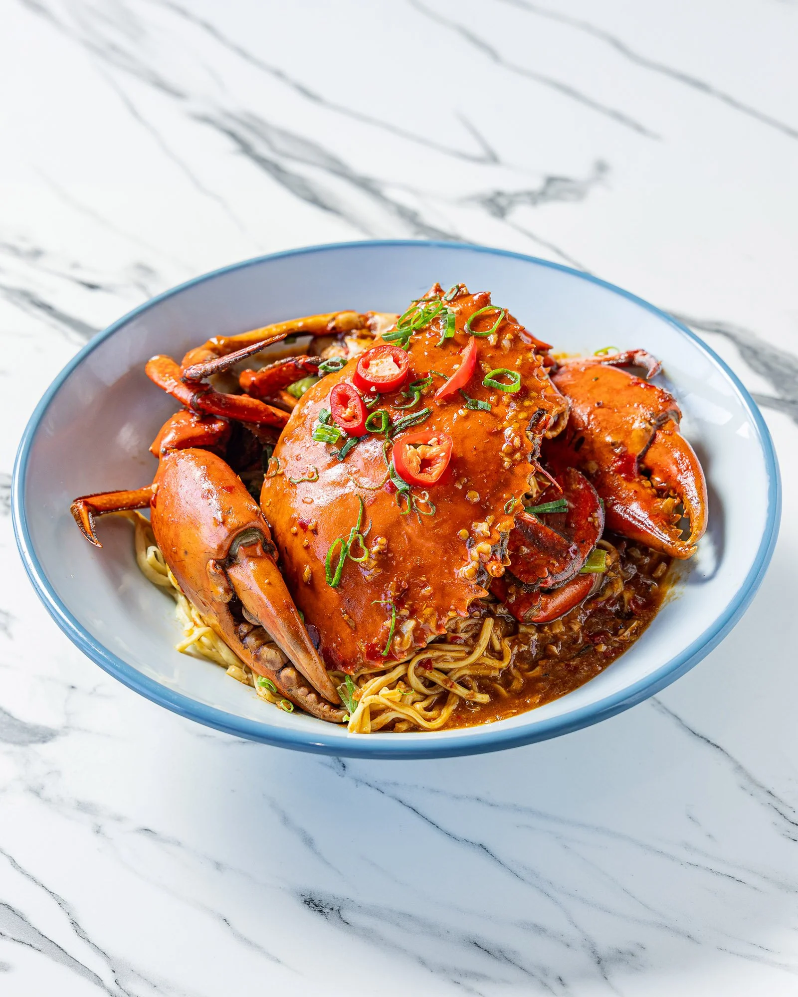 A bowl of seafood noodle soup with lobster, garnished with sliced green onions and red chili peppers, on a marble surface.