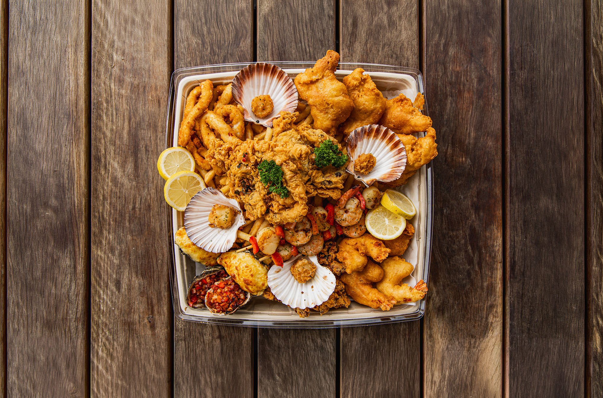 A seafood platter with fried fish, shellfish, lemon slices, and garnishes on a tray placed on a wooden table.
