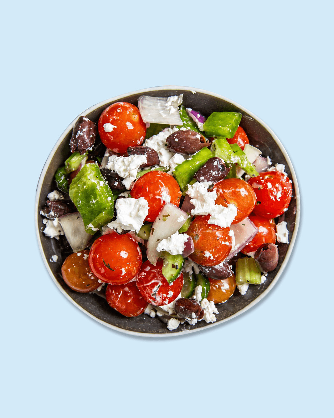 A bowl filled with cherry tomatoes, green bell pepper, onion, olives, feta cheese, and herbs on branded paper background.