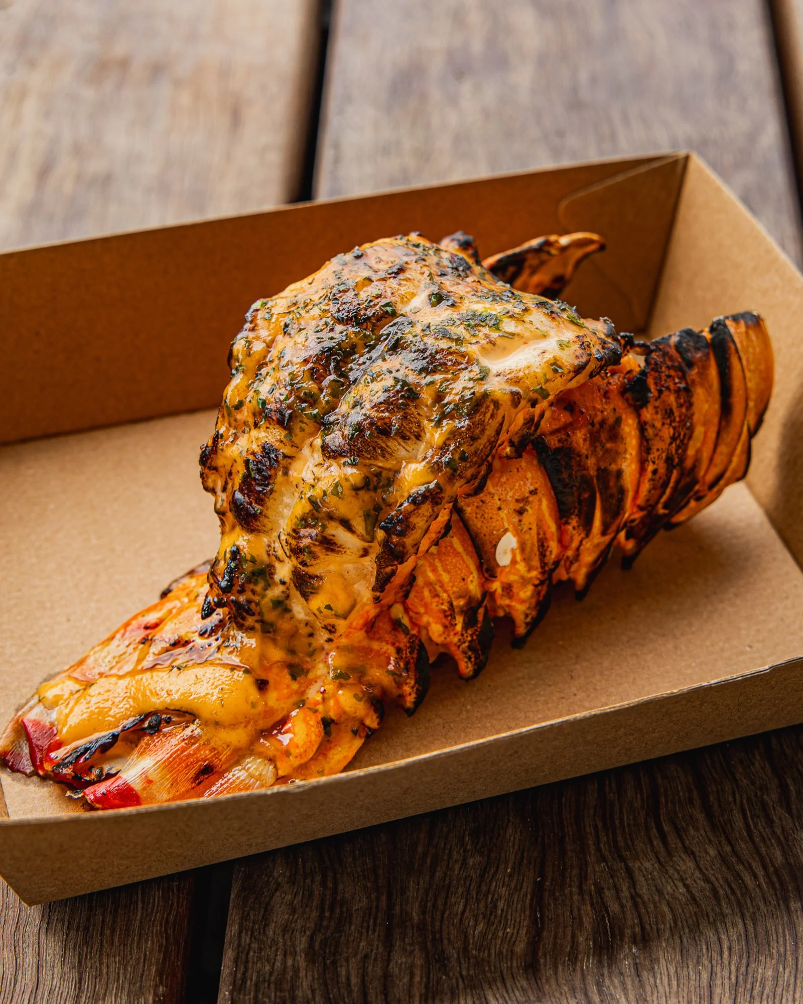 A cooked lobster with melted cheese and herbs in a brown cardboard takeout box, placed on a dark wooden surface.