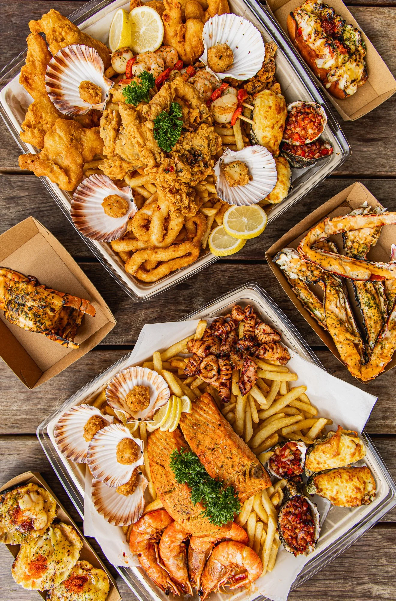 Assortment of seafood dishes including fried fish, shrimp, crawfish, and sushi, with lemon wedges, French fries, and various dipping sauces on a wooden table.