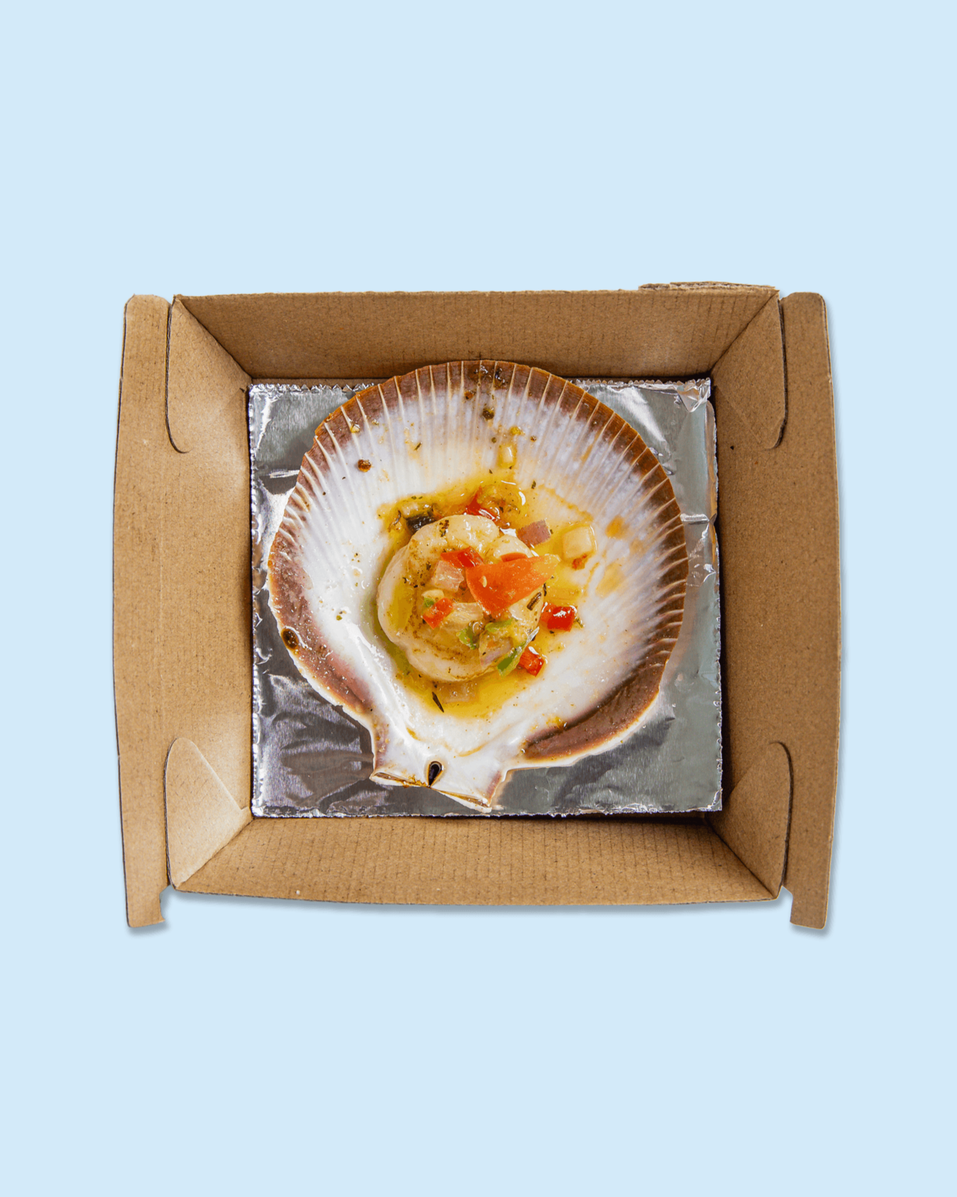A partially eaten scallop served in its shell with chopped vegetables and sauce, placed in a cardboard takeout container lined with aluminum foil.