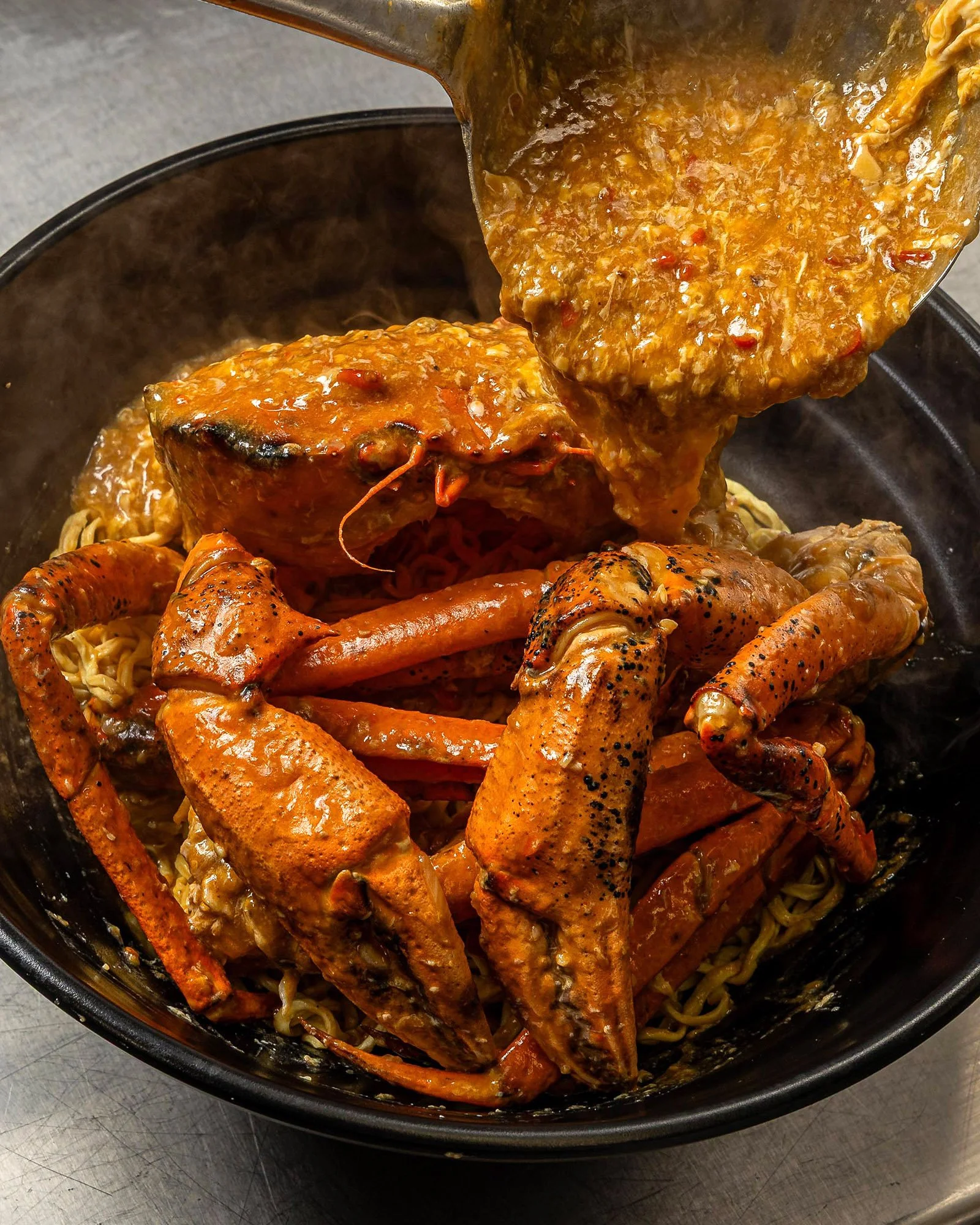 Lobster tail and skewered shrimp on top of ramen noodles with thick sauce being poured into a black container.