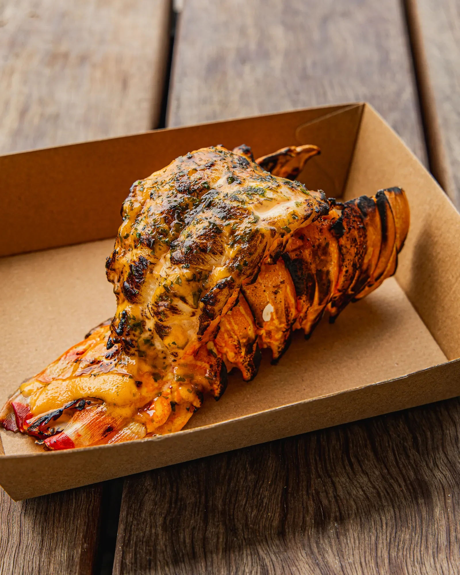 Grilled lobster tail with melted cheese and herbs, served on a brown cardboard tray on a wooden table.
