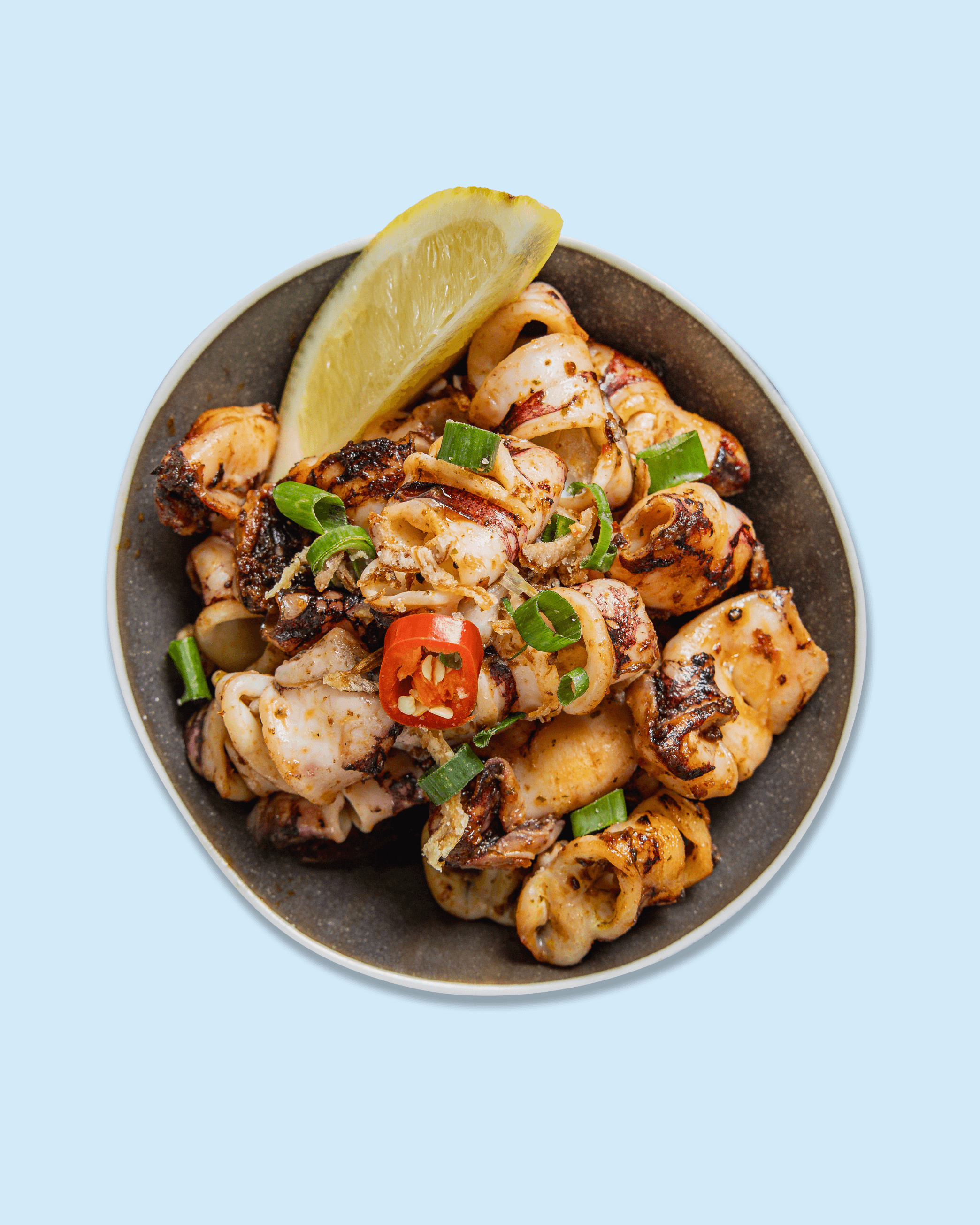 A bowl of grilled seafood including squid and octopus, garnished with sliced green onions and red chili peppers, on a paper with "Nicholas catering & events" branding.