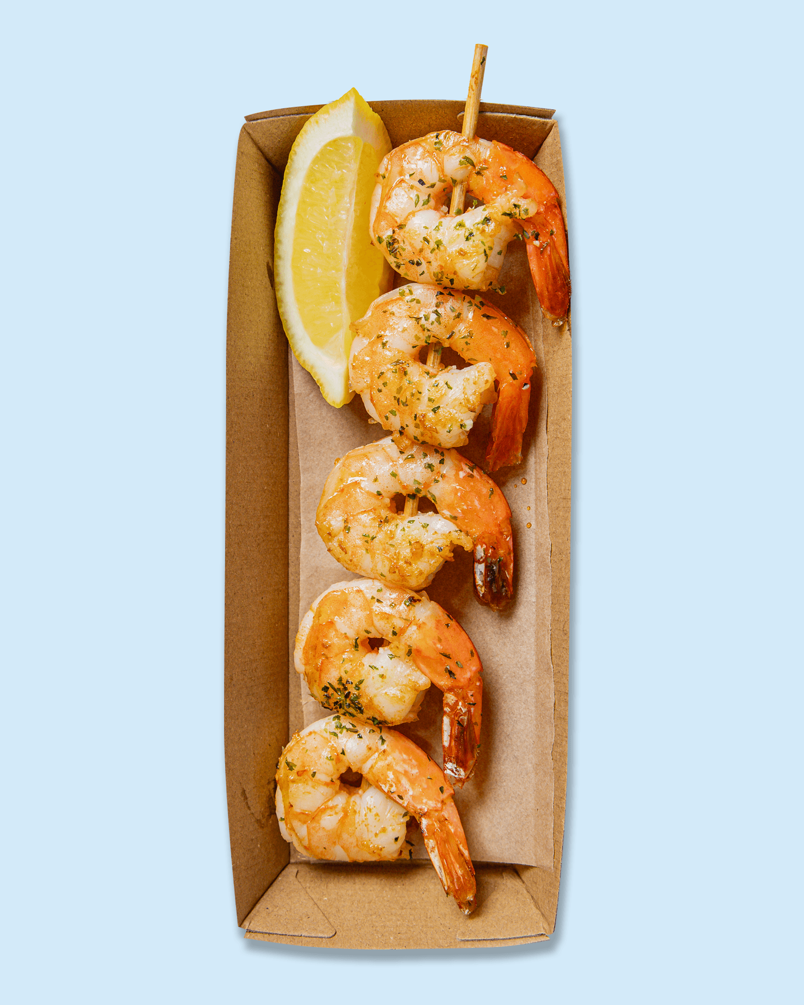 A cardboard serving tray with cooked shrimp garnished with herbs, lemon wedges, and a small wooden skewer, on printed paper with a logo and text.