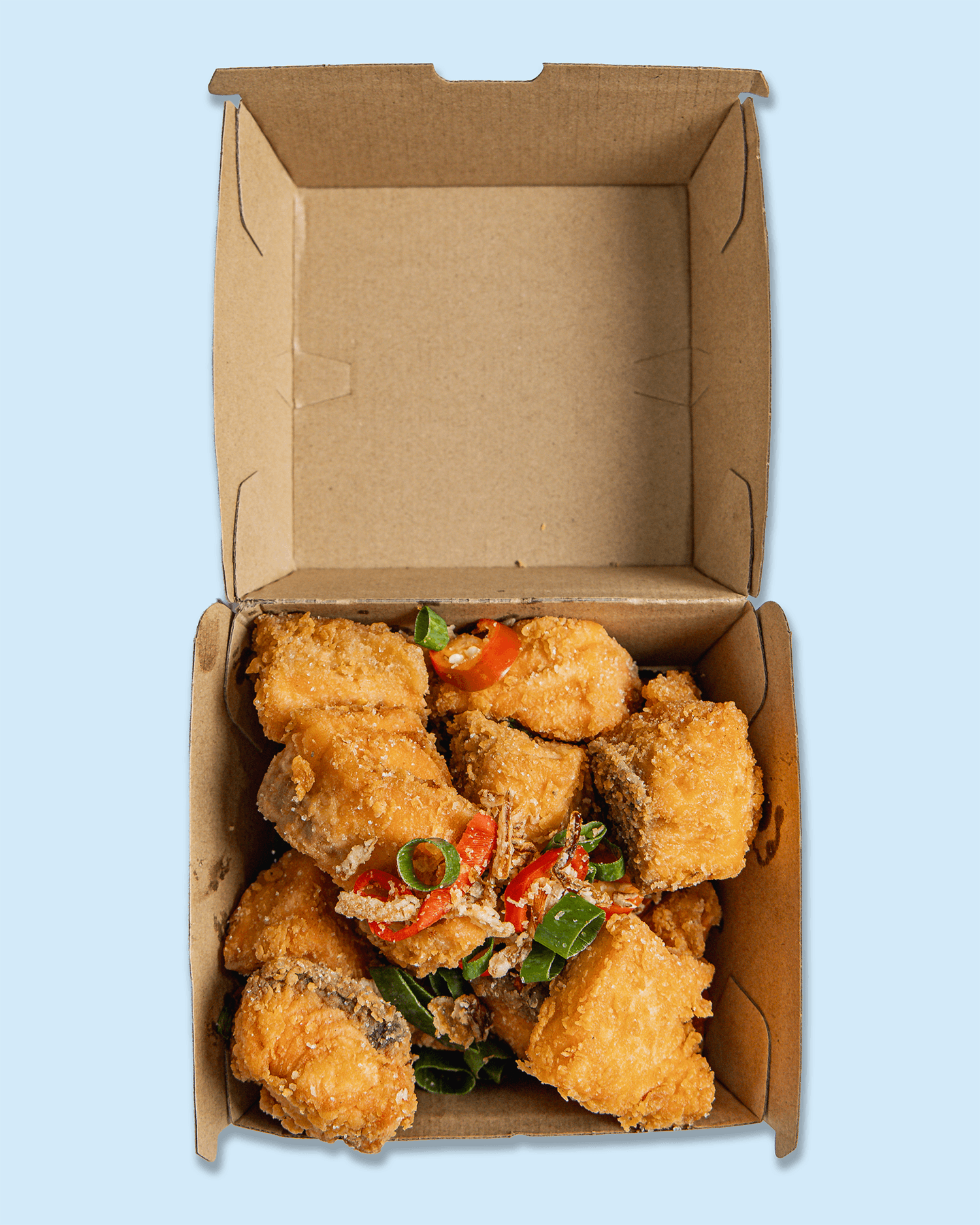 Fried chicken pieces topped with chopped green onions and red chili peppers inside a cardboard takeout box on a branded paper surface.