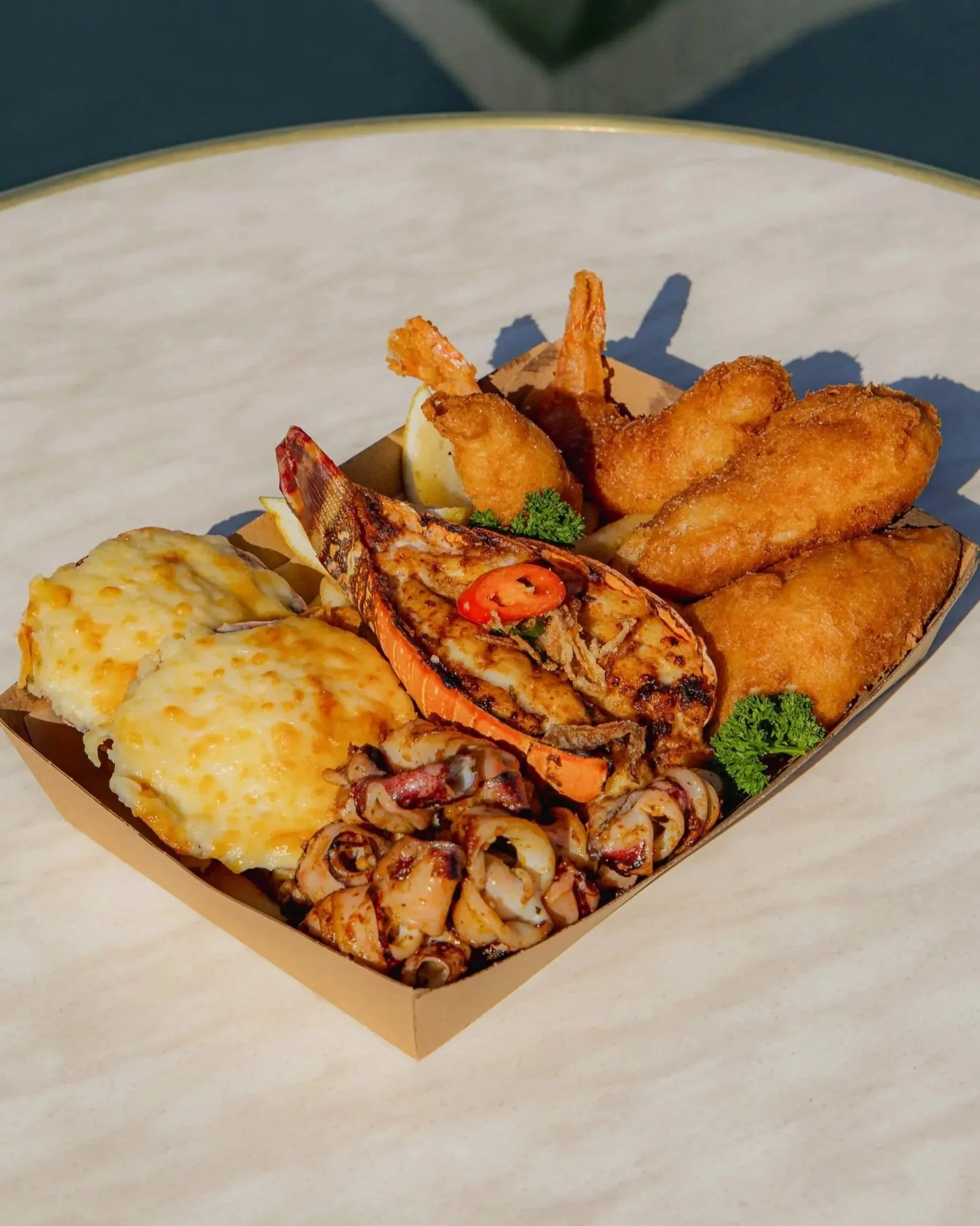 A box of seafood with fried shrimp, battered fish, scalloped potatoes, fried octopus, and French fries on a marble surface.