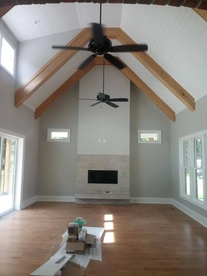 Empty living room with hardwood floors, a fireplace, large windows, and a vaulted ceiling with wooden beams and two ceiling fans.