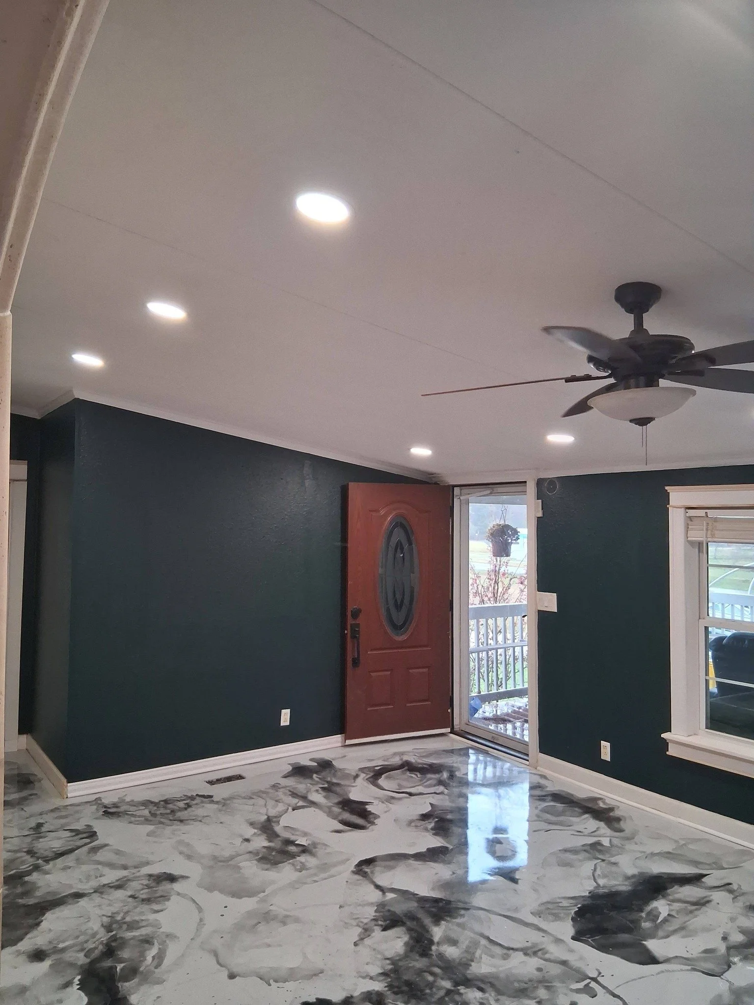 Interior view of a living room with dark green walls, white ceiling with recessed lights, a ceiling fan, a wooden front door, a window with white trim, and a marble floor with black and white patterns.