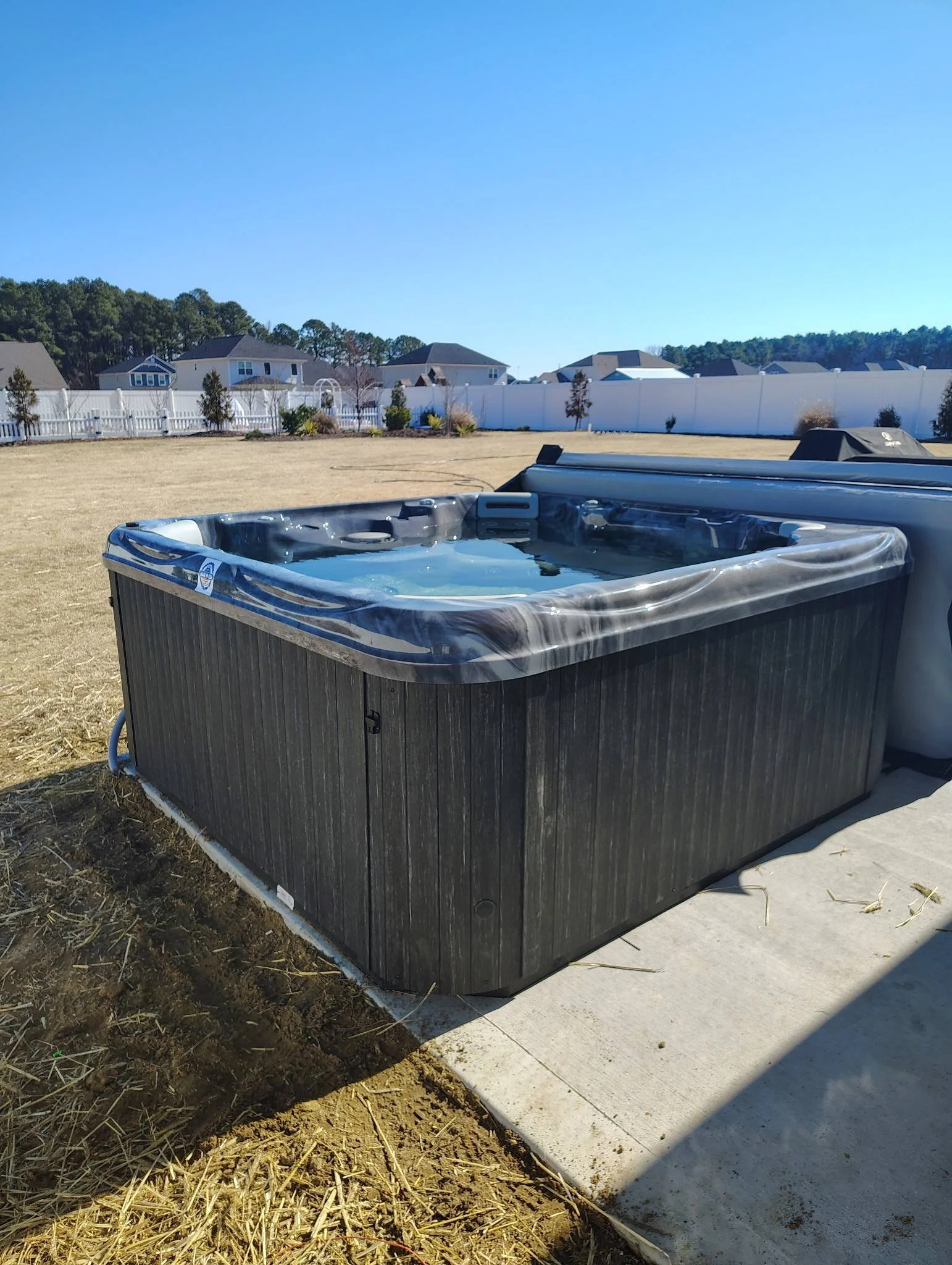 Hot tub filled with water on a concrete pad in a backyard with a grass area and houses in the background under a clear blue sky.