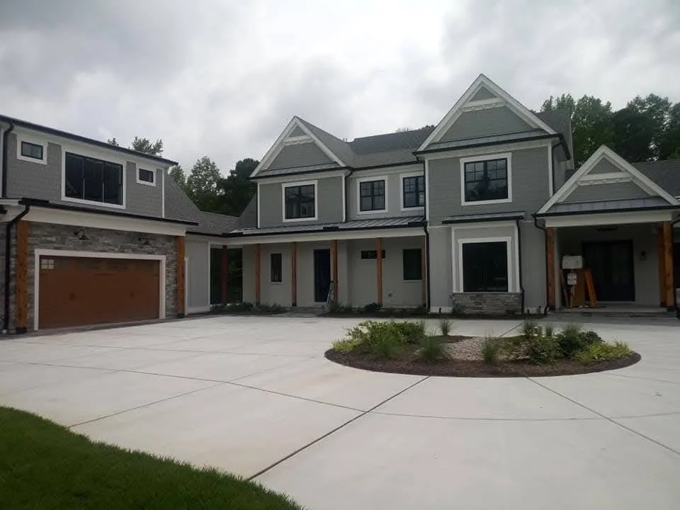 Newly built two-story house with a large driveway, gray exterior, and wooden accents, surrounded by landscaping plants.
