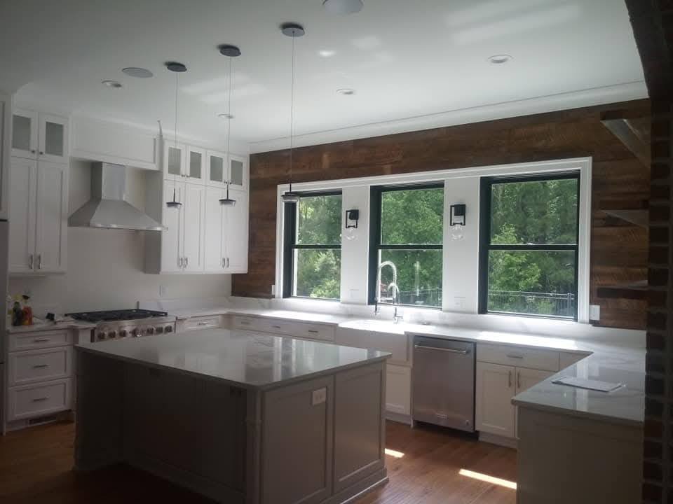 A modern kitchen with white cabinets, a kitchen island, a stainless steel dishwasher, a stove with an overhead vent, and three large windows overlooking greenery. There are pendant lights hanging above the island and two wall sconces on the wall.