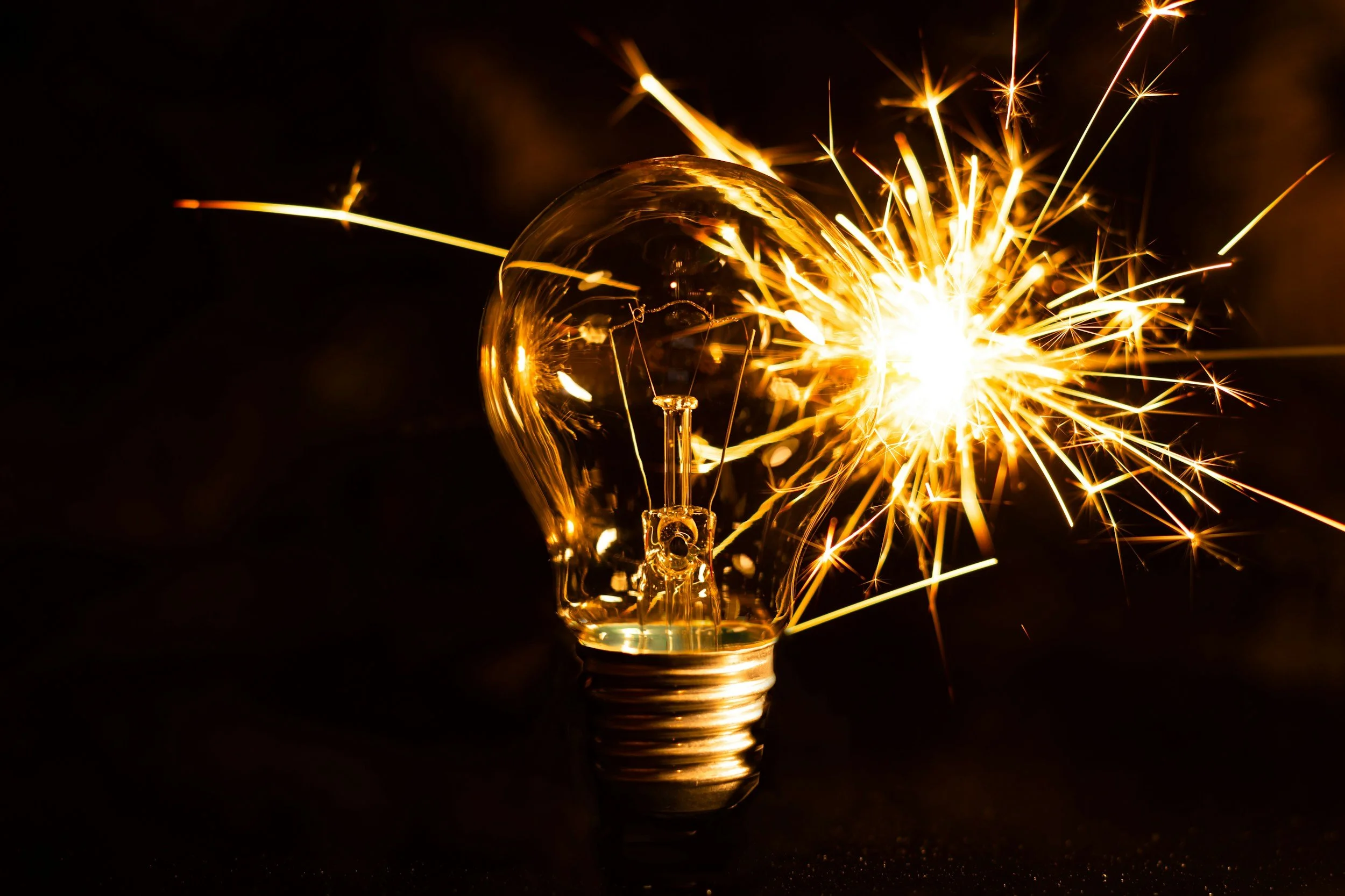 A close-up of a clear light bulb with a sparkler igniting next to it, emitting bright sparks against a dark background.