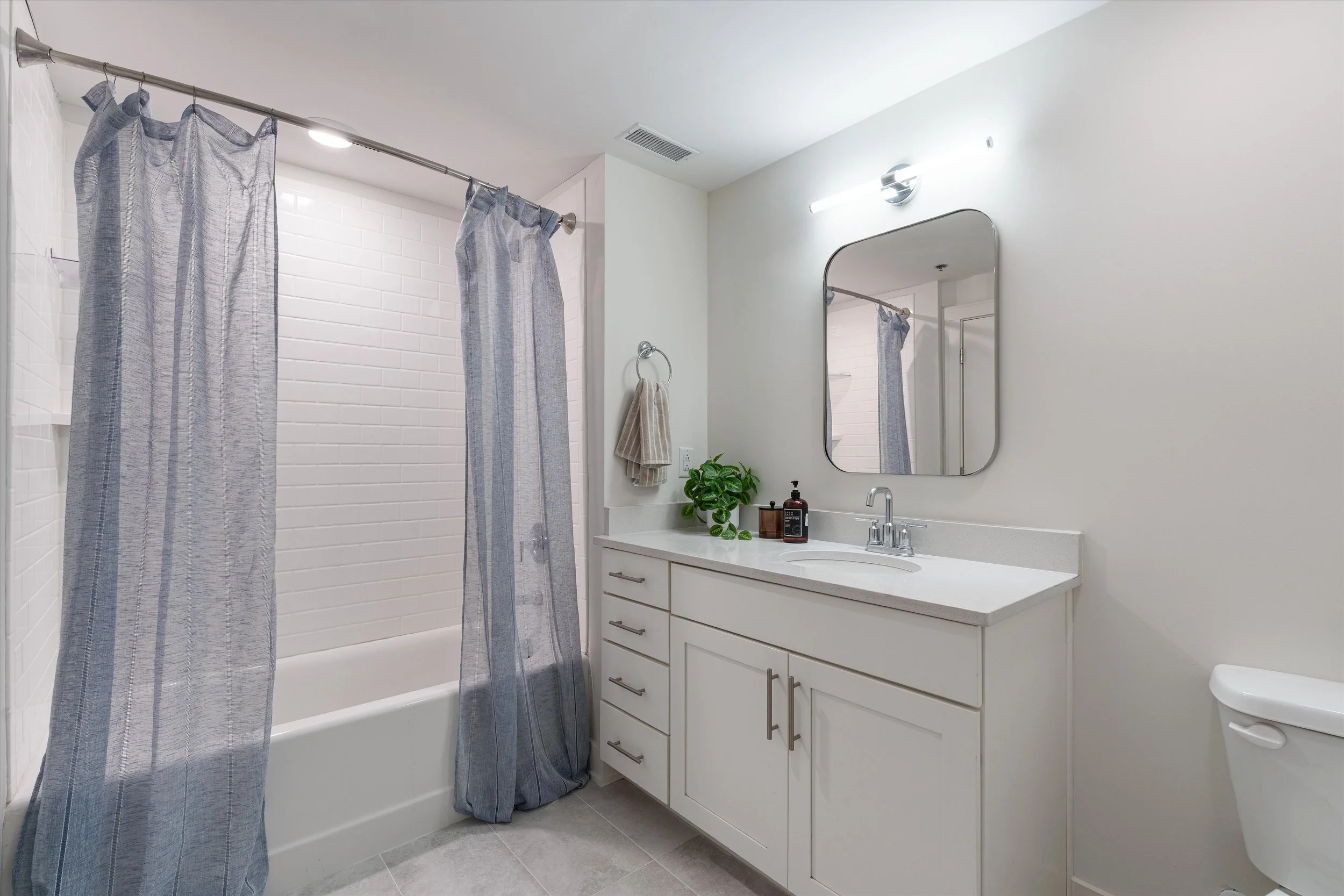 Modern bathroom with a shower/tub combination, blue striped shower curtains, white tiled wall, white vanity with a mirror, countertop with a small plant and soap dispenser, and a toilet.