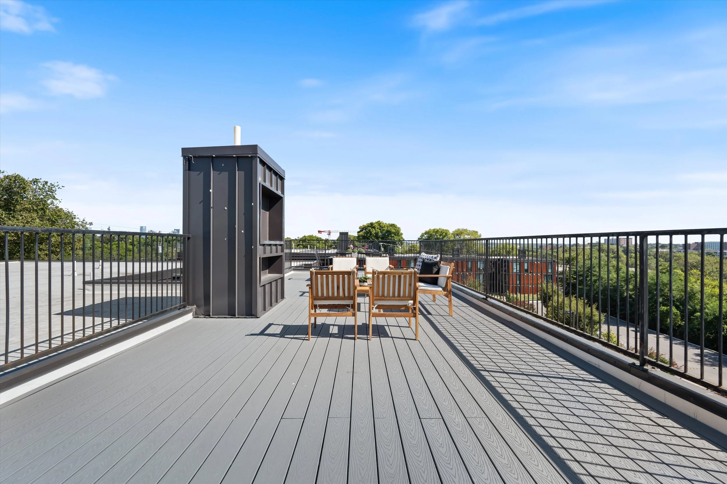 Rooftop terrace with outdoor seating, wooden chairs, black railing, and a black storage unit on a sunny day with blue sky.