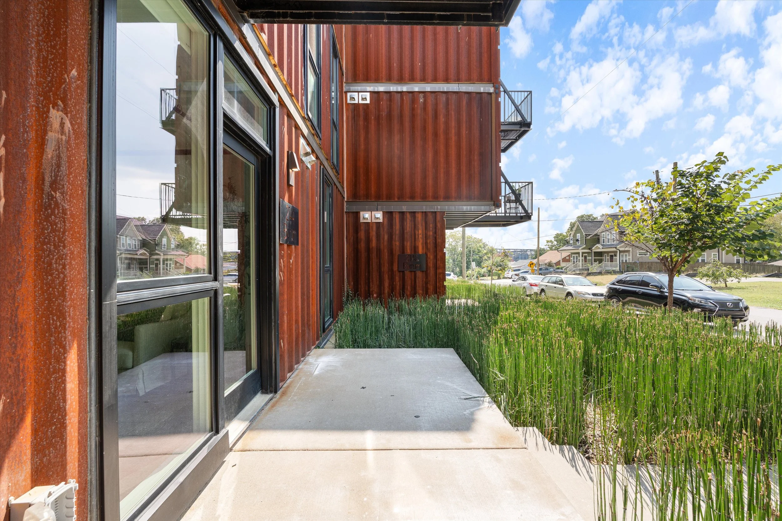 View of a modern building's exterior with rust-colored metal siding, large glass sliding door, small concrete patio, green grass, trees, parked cars, and houses in the background.