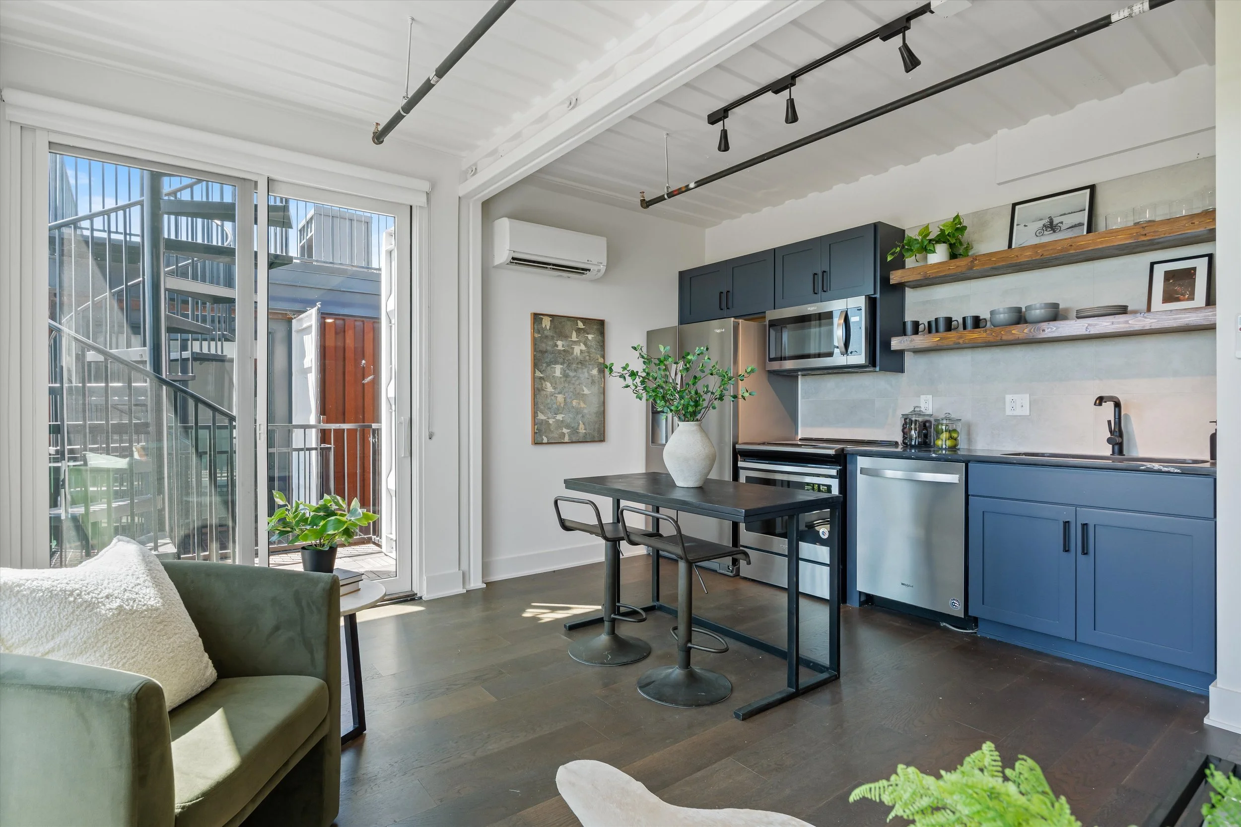 Open-concept kitchen and living area with dark blue cabinets, stainless steel appliances, floating wooden shelves, black track lighting, dark hardwood floors, and sliding glass door leading to balcony with outdoor stairs visible.