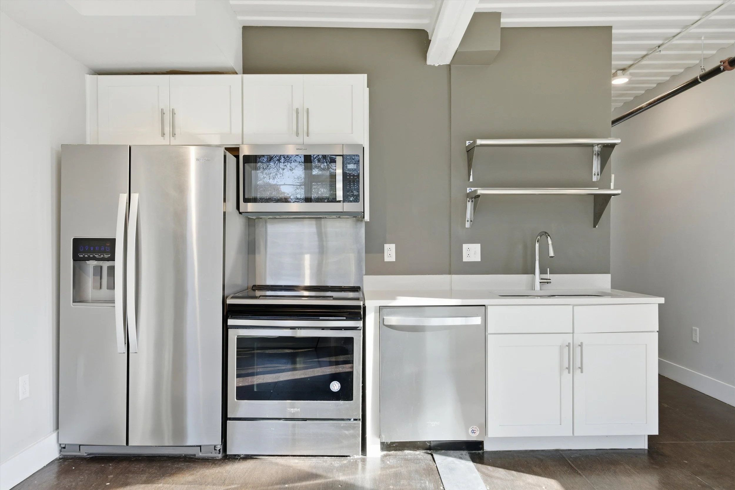 Stainless steel refrigerator, microwave, oven, and dishwasher in a modern kitchen with white cabinets and gray wall, with open shelves above the sink.
