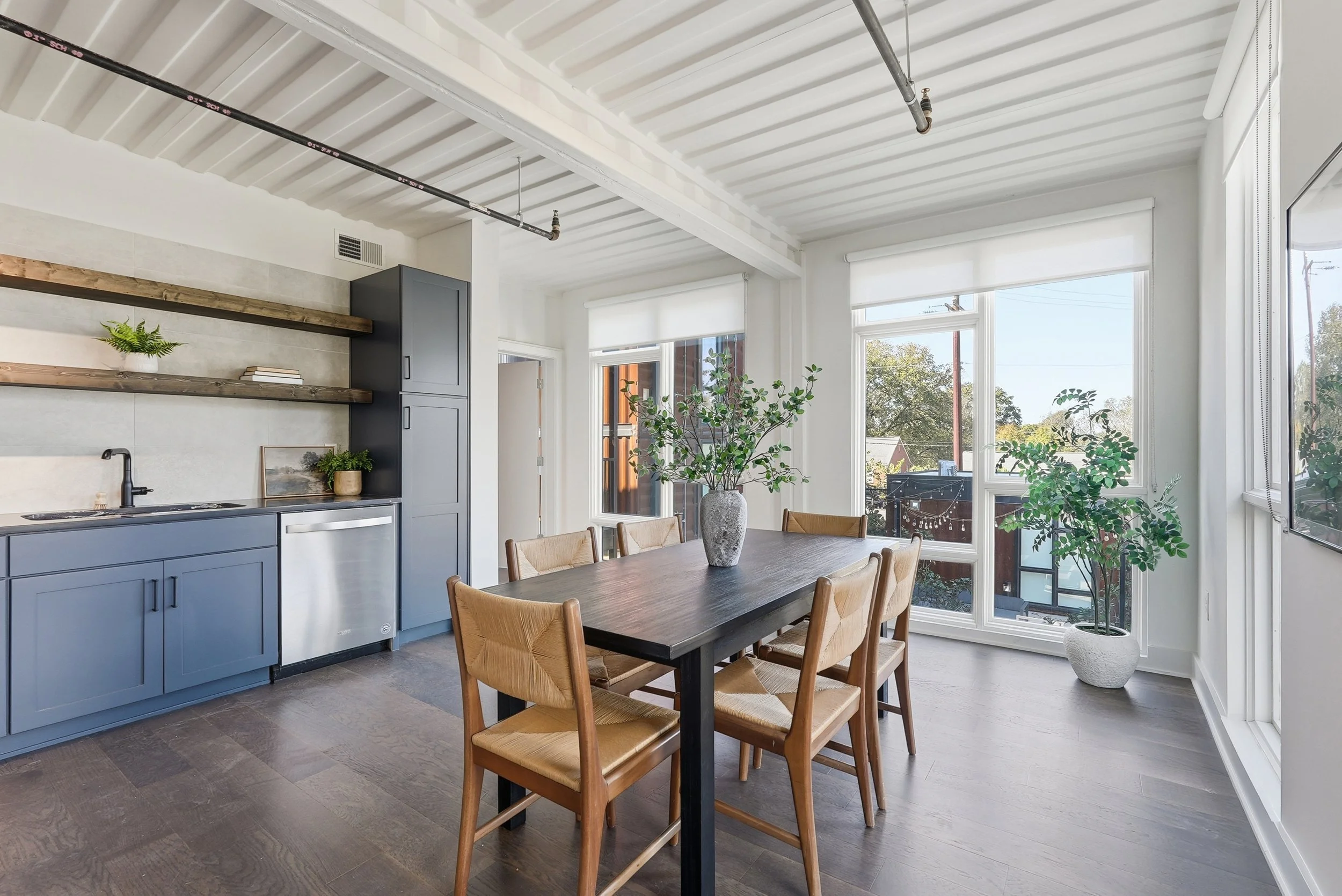 Bright dining area with a wooden table, six wooden chairs with woven seats, large windows with white blinds, a potted plant, and a minimalist kitchen with blue cabinets and open wooden shelves.