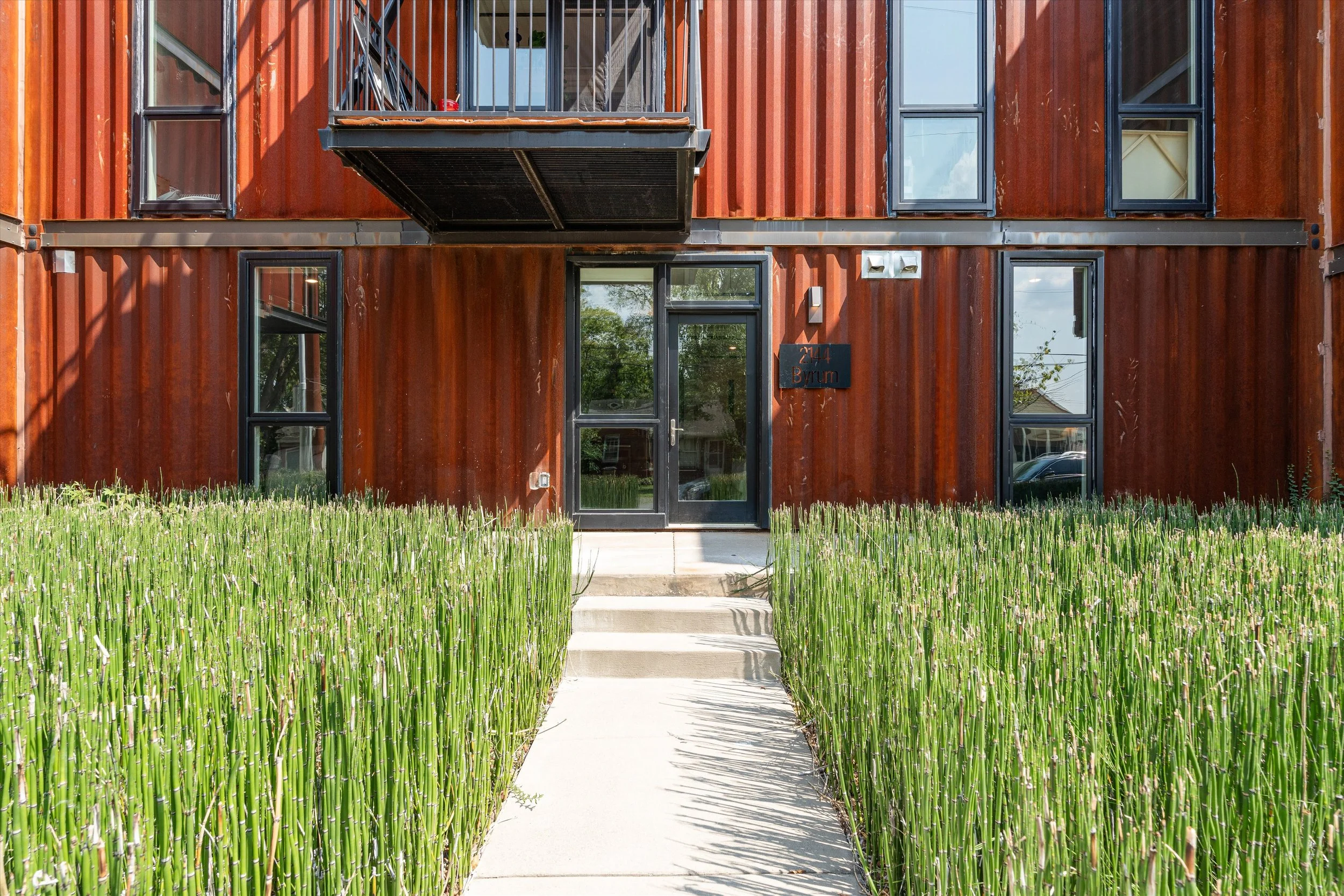 Front view of a modern residential building with a rusty metal exterior, black-framed windows, and a glass door, with a concrete pathway and tall green plants on either side leading to the entrance.