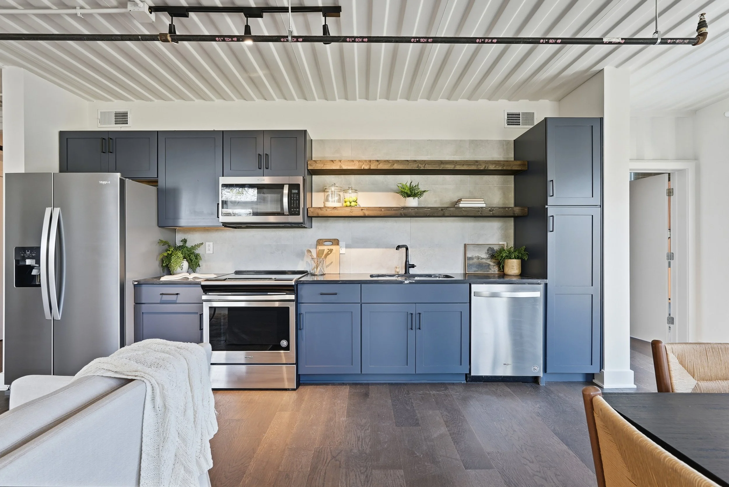 Modern kitchen with dark blue cabinets, stainless steel appliances, open wooden shelves, and potted plants.