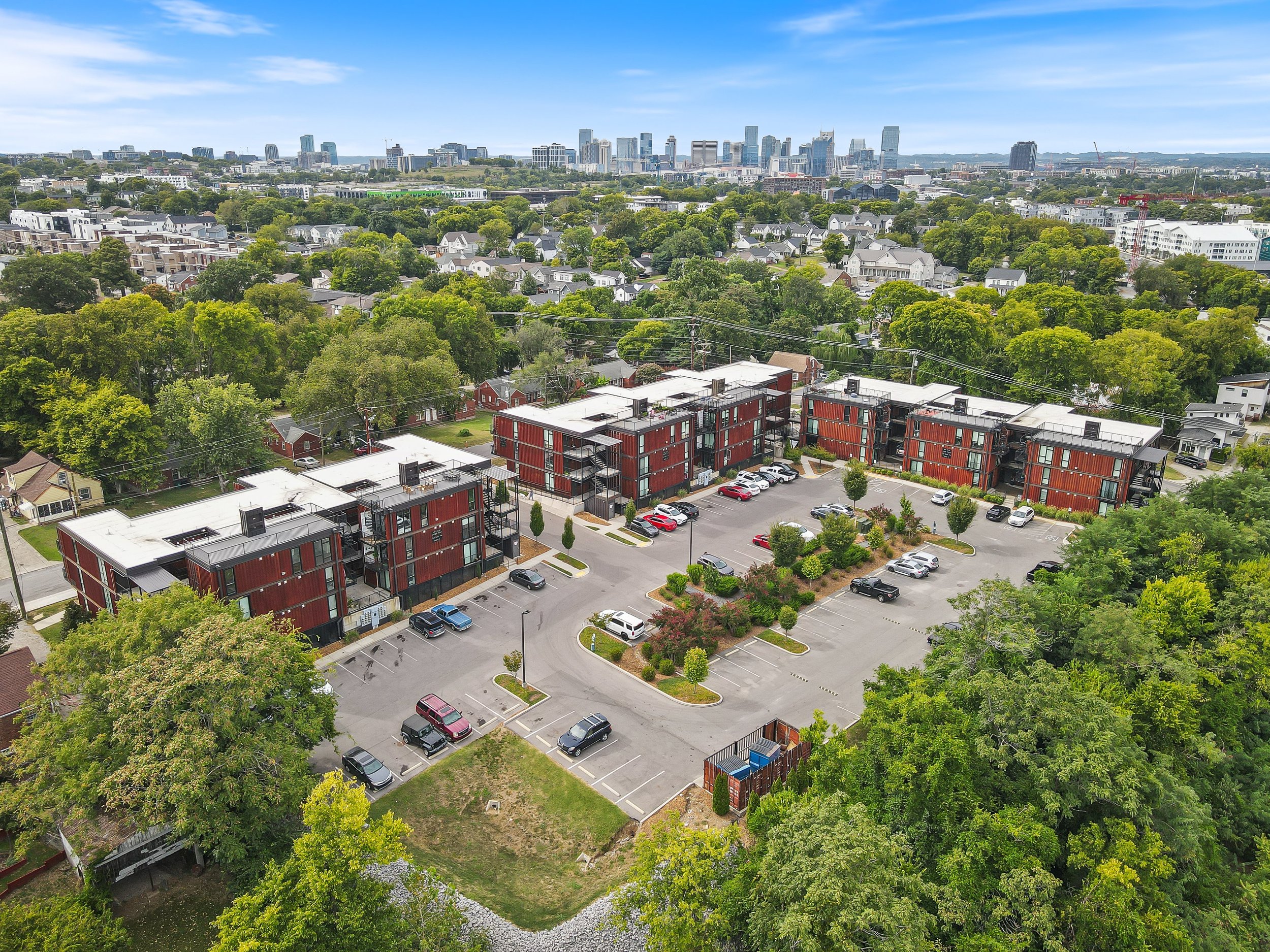 Aerial view of a residential apartment complex with multiple buildings, parking lots, surrounded by greenery, with a city skyline in the background under a blue sky.