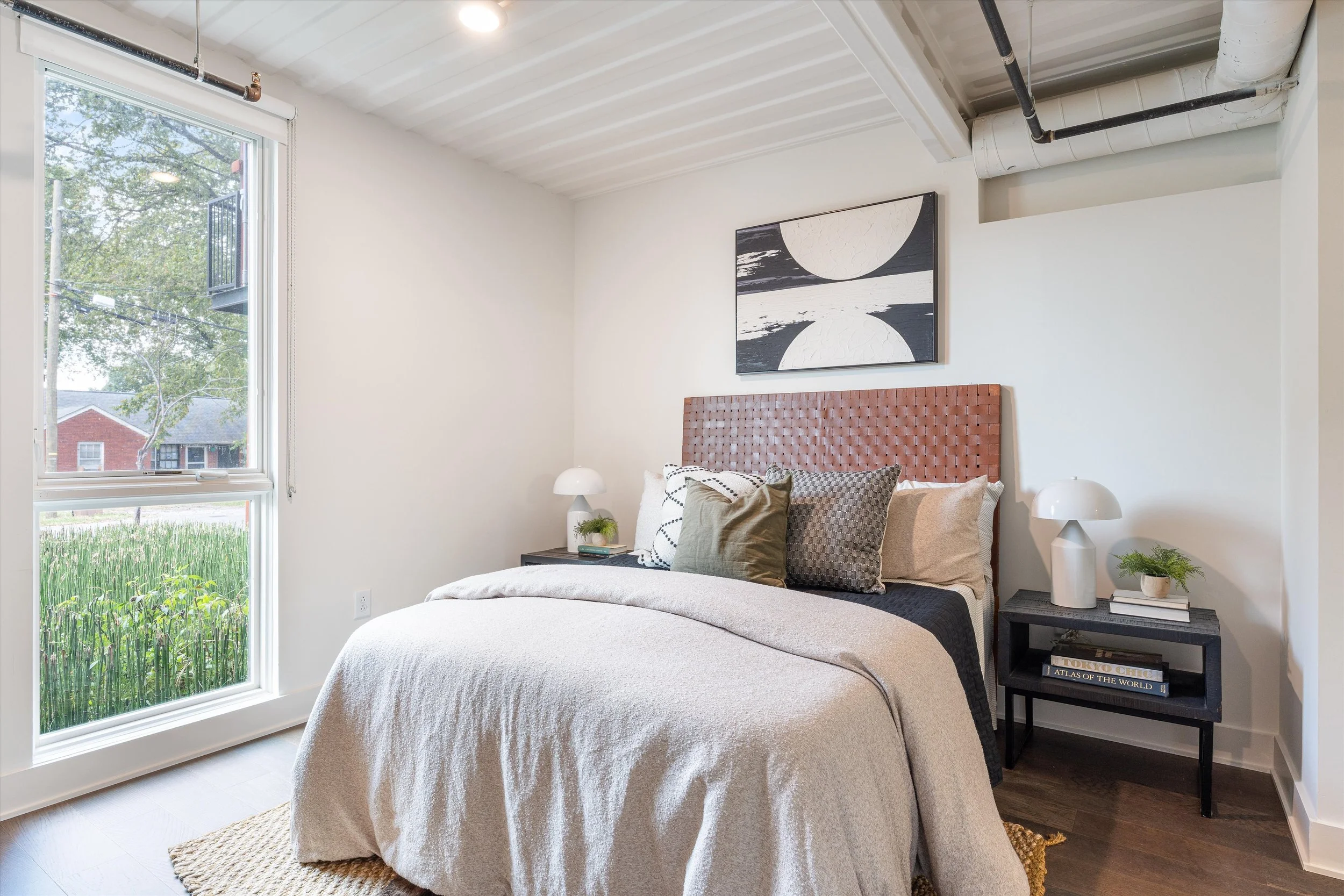 Bedroom with a large bed, brown woven headboard, white bedding, and multiple pillows. Two black bedside tables with white lamps, a window showing trees and a red brick building outside, and a black and white abstract art piece above the bed.