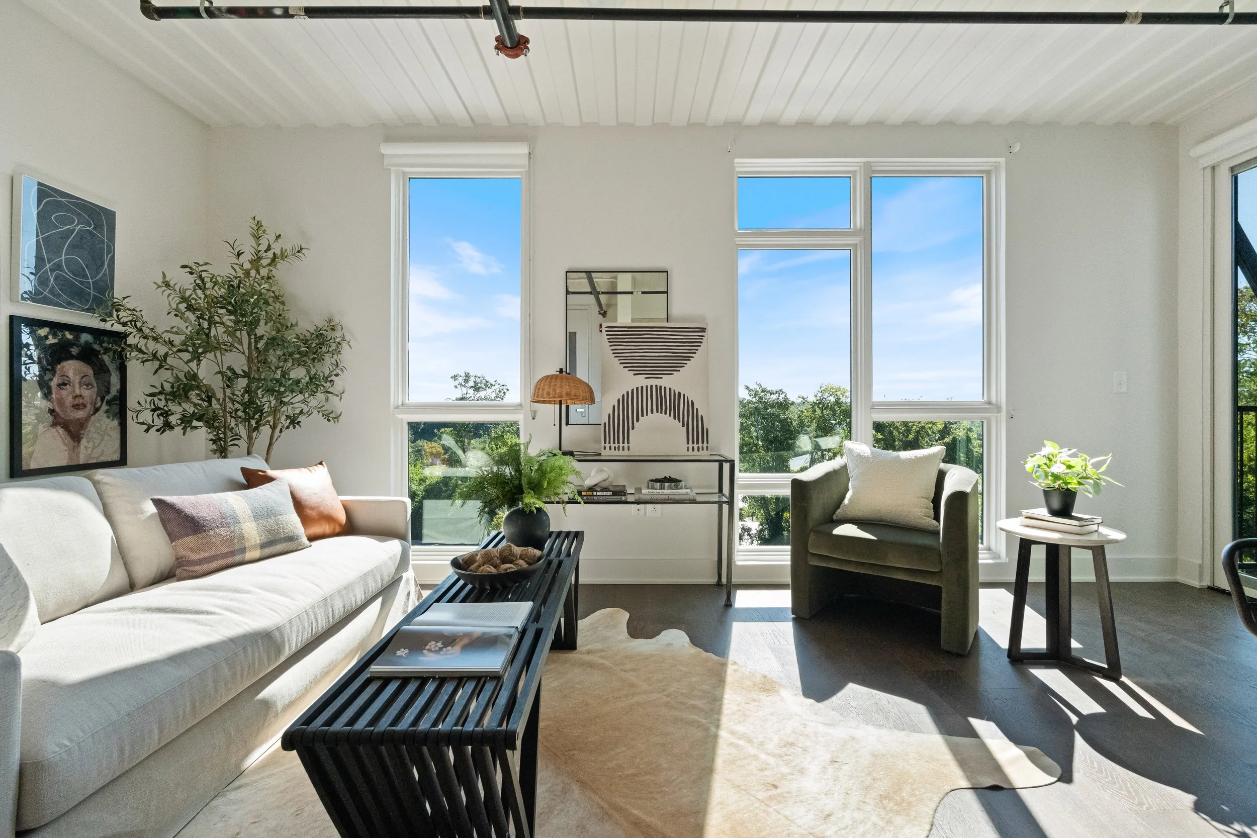Bright living room with large windows, white walls, a white sofa with throw pillows, a black coffee table, a gray armchair with a pillow, and houseplants on side tables, featuring contemporary decor and artwork.