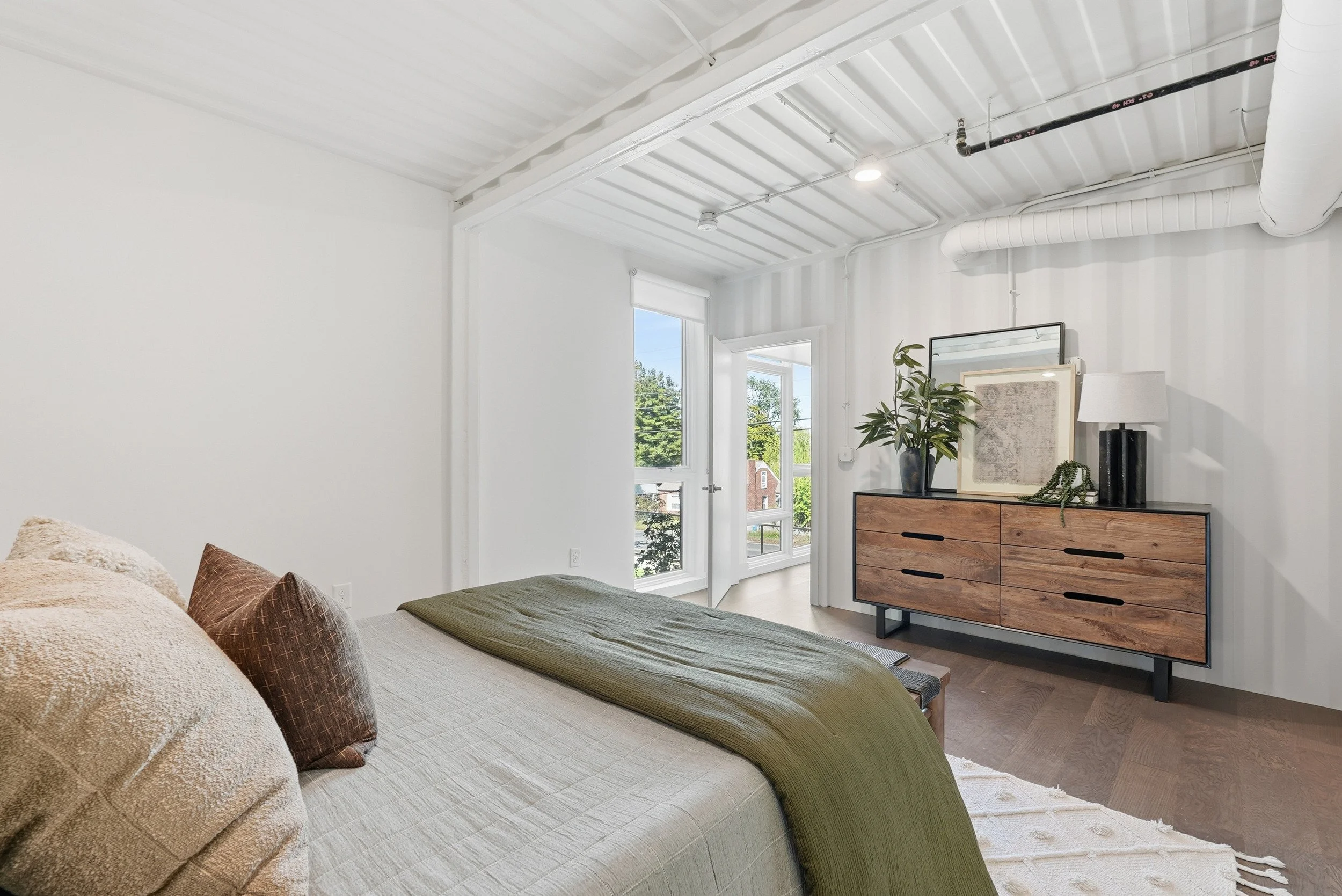 Bright bedroom with white walls, a bed with pillows and green throw, a wooden and black dresser with decorative items, and large windows showing outdoor trees.