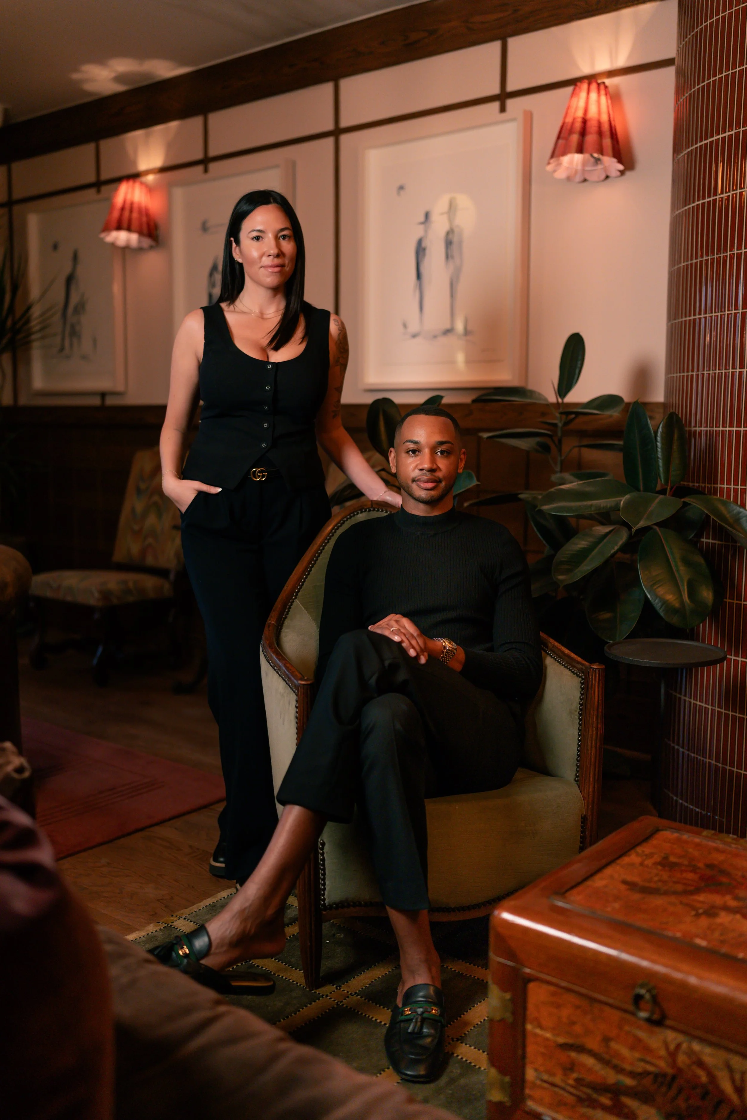 A woman standing next to a seated woman in a cozy, stylish living room with artwork on the wall, plants, and warm lighting.