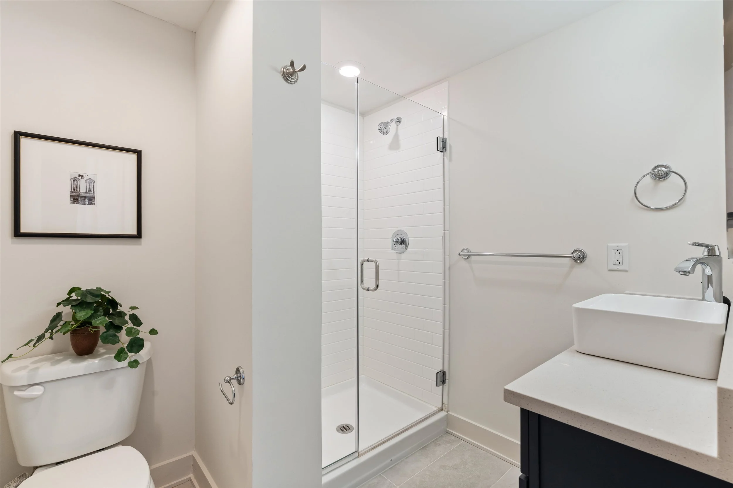 A modern bathroom with a glass shower enclosure, a white rectangular vessel sink on a grey vanity, a towel bar, a wall-mounted mirror, a framed black-and-white photograph, and a small potted plant on top of the toilet tank.