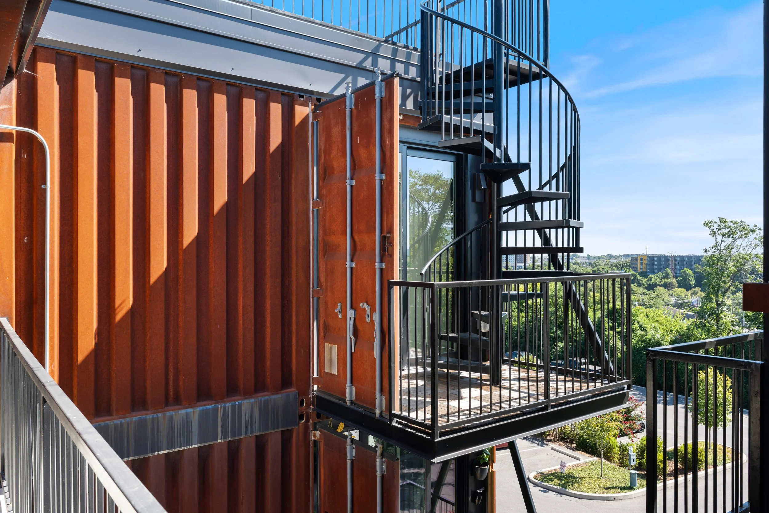 Exterior view of a modern building with a black spiral staircase on a balcony, metal railings, a rust-colored metal wall, large glass windows, and a cityscape with trees and buildings in the background under a clear blue sky.
