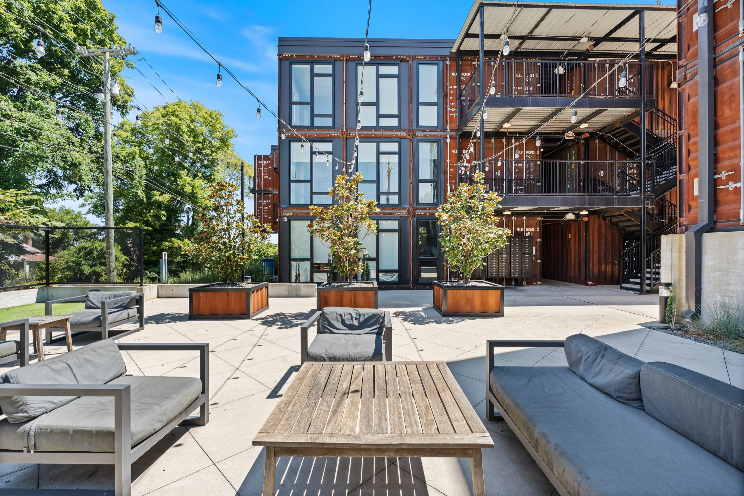 Outdoor patio with modern furniture, potted plants, and string lights, enclosed by a building made of stacked shipping containers.