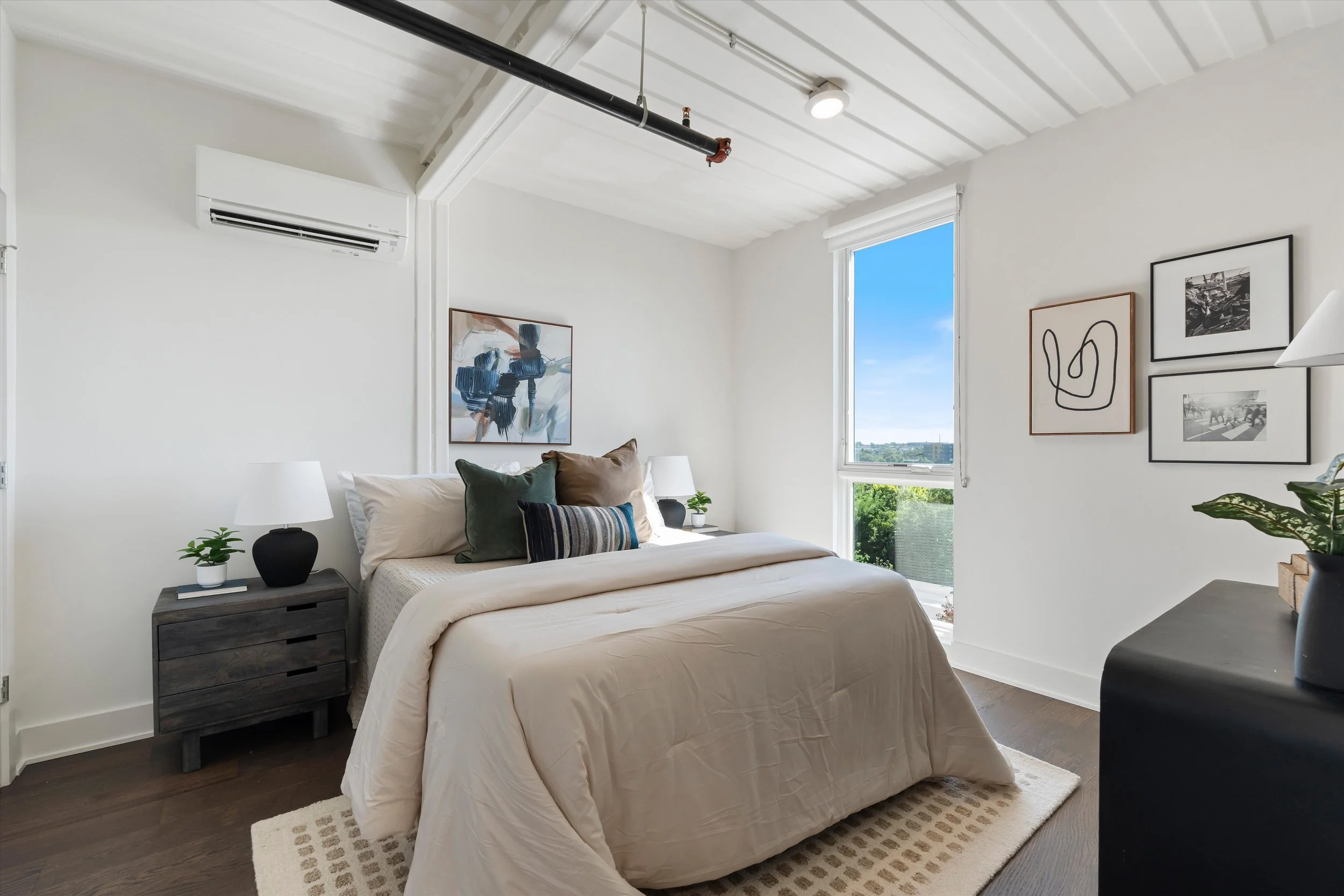 Bedroom with a white bed, dark wood nightstand, and framed art on the walls. Large window shows blue sky and greenery outside.