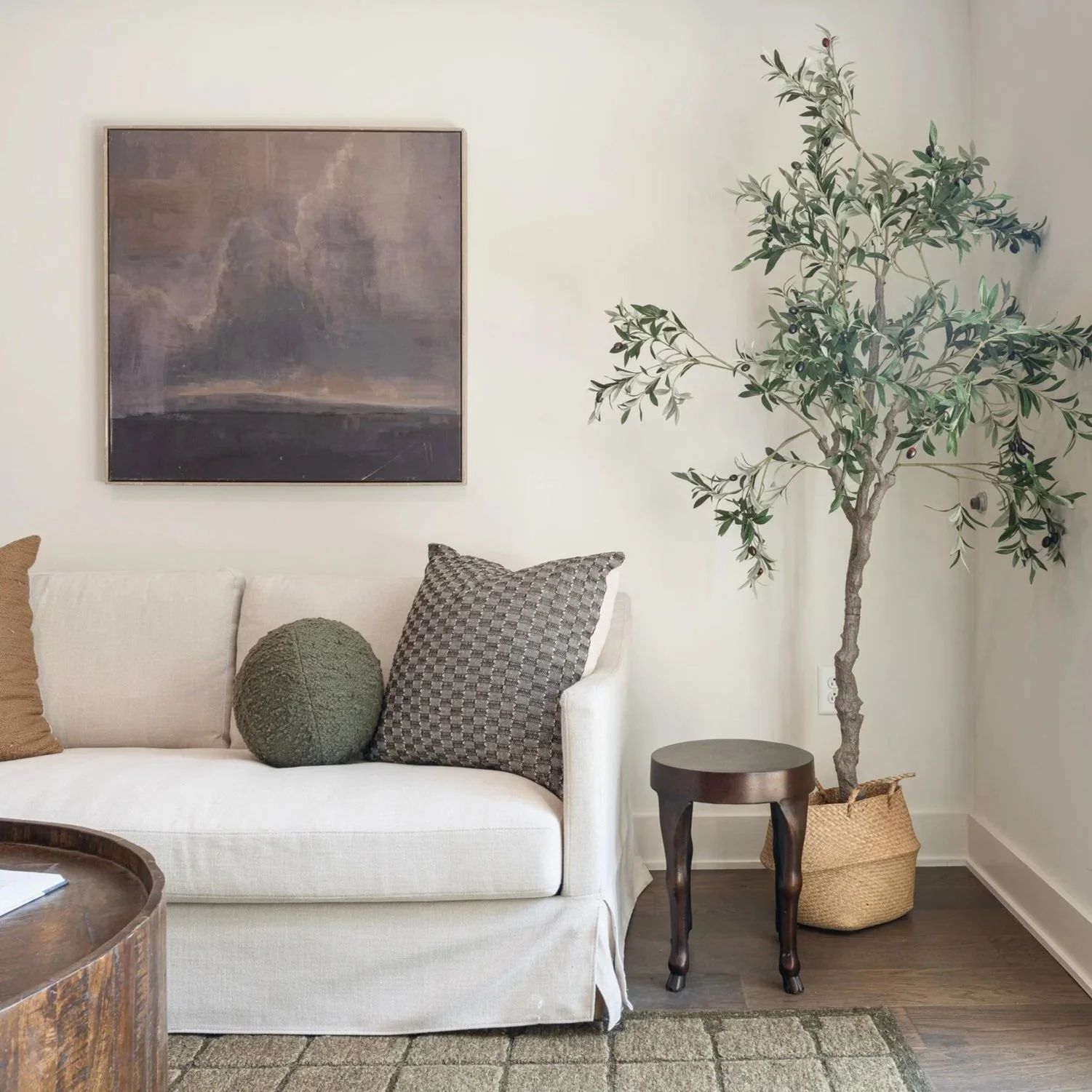 Living room with white sofa, decorative pillows, landscape painting, small dark wooden table, potted olive tree in a basket, and rug on hardwood floor.