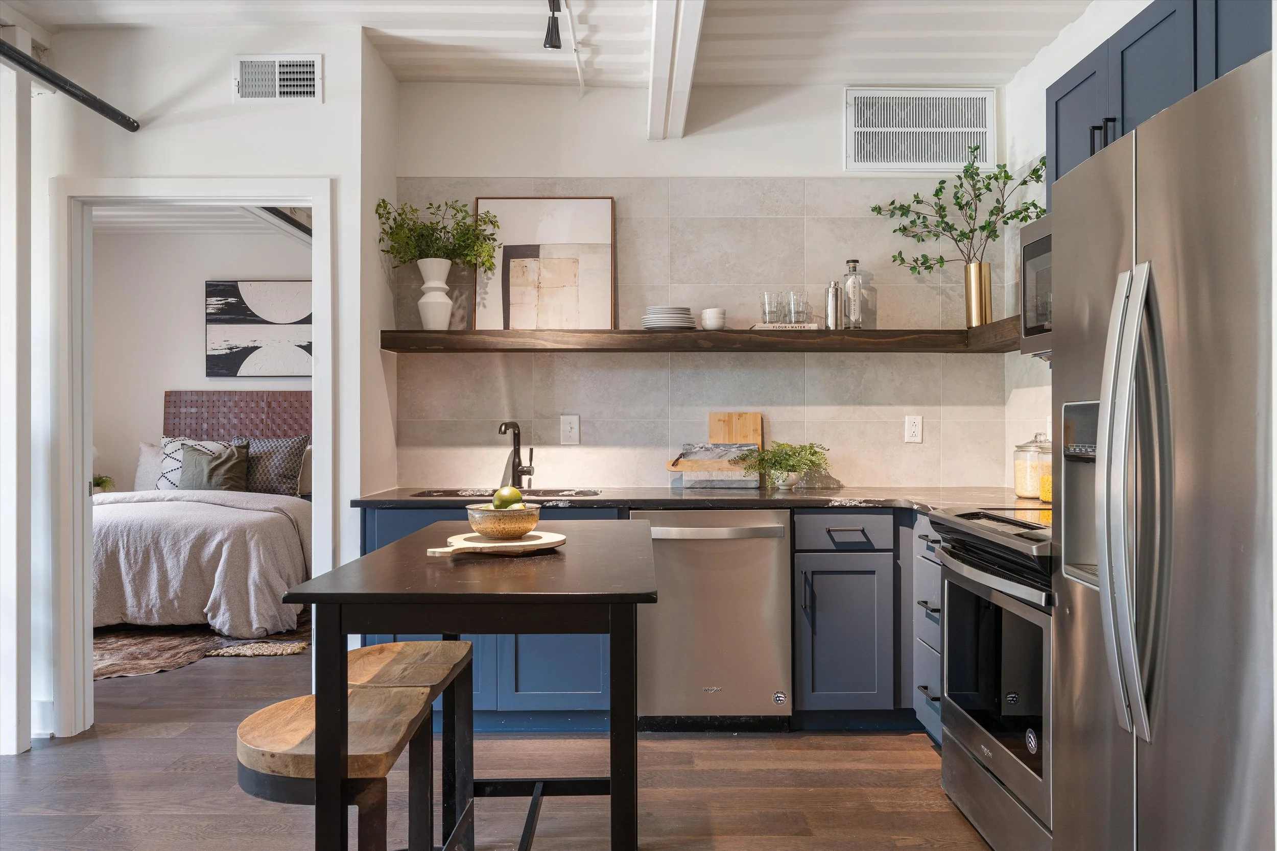 Modern kitchen with blue cabinets, stainless steel appliances, black countertop, and open wooden shelf with decor. Part of living space with bedroom visible in background.