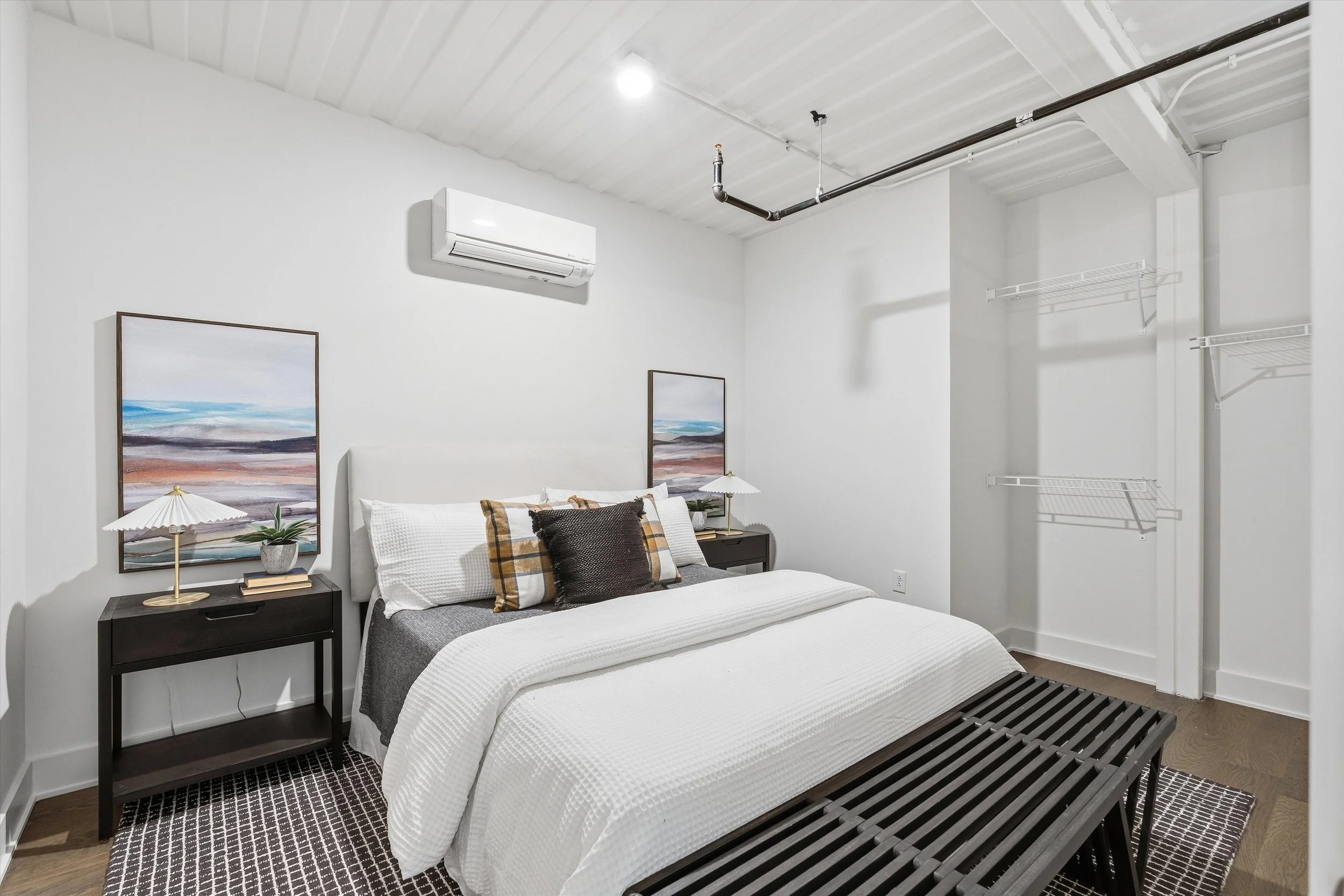 Bedroom with white walls, a bed with white bedding and decorative pillows, two black nightstands with lamps, two landscape paintings above the bed, an air conditioning unit, and an open closet with wire shelves.